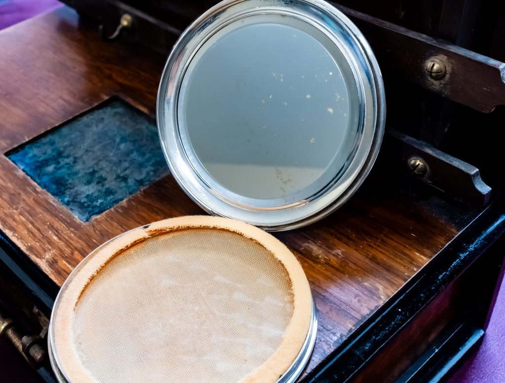 A vintage-style round compact mirror with a silver rim resting on a dark wooden box, next to an open round case with a beige textured insert.