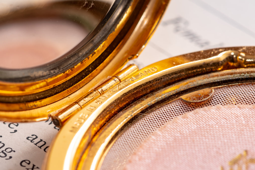 Close-up of an open vintage gold compact case with a pink pressed powder pan visible inside, resting on printed paper.