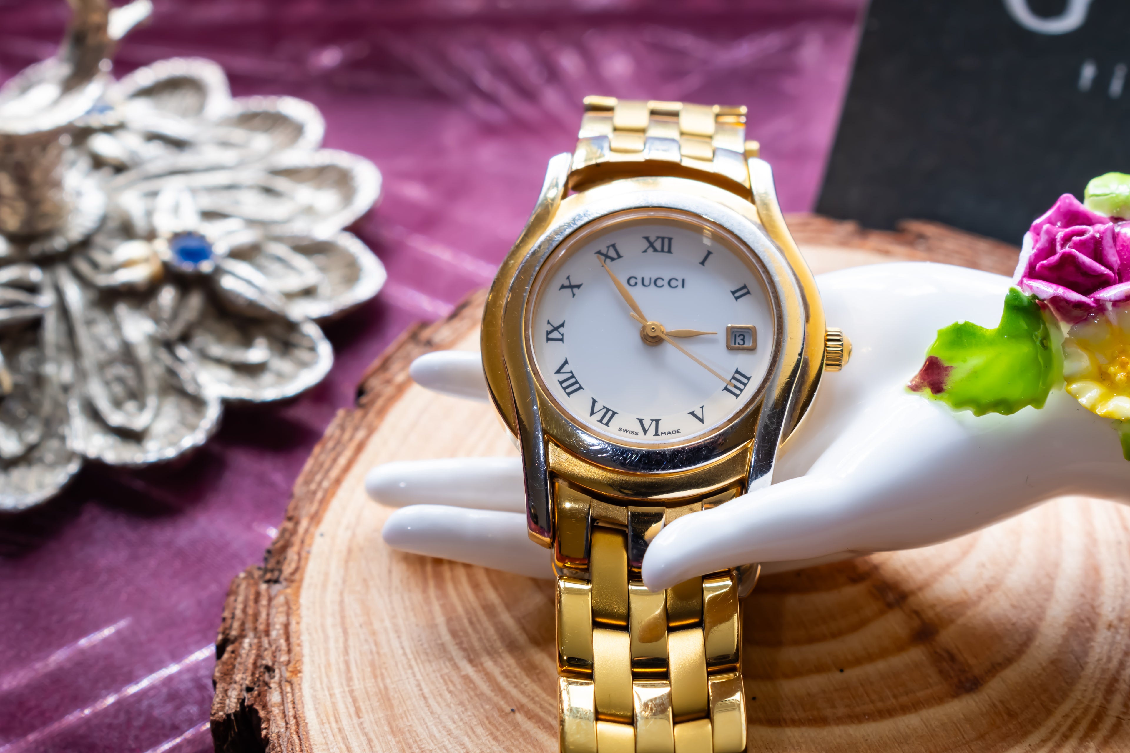 A gold-tone Gucci watch with a white dial and Roman numerals, displayed on a white mannequin hand resting on a wood slice over a purple fabric background.