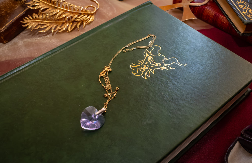 A gold chain necklace with a faceted purple crystal pendant resting on a dark green book with gold foil detailing.
