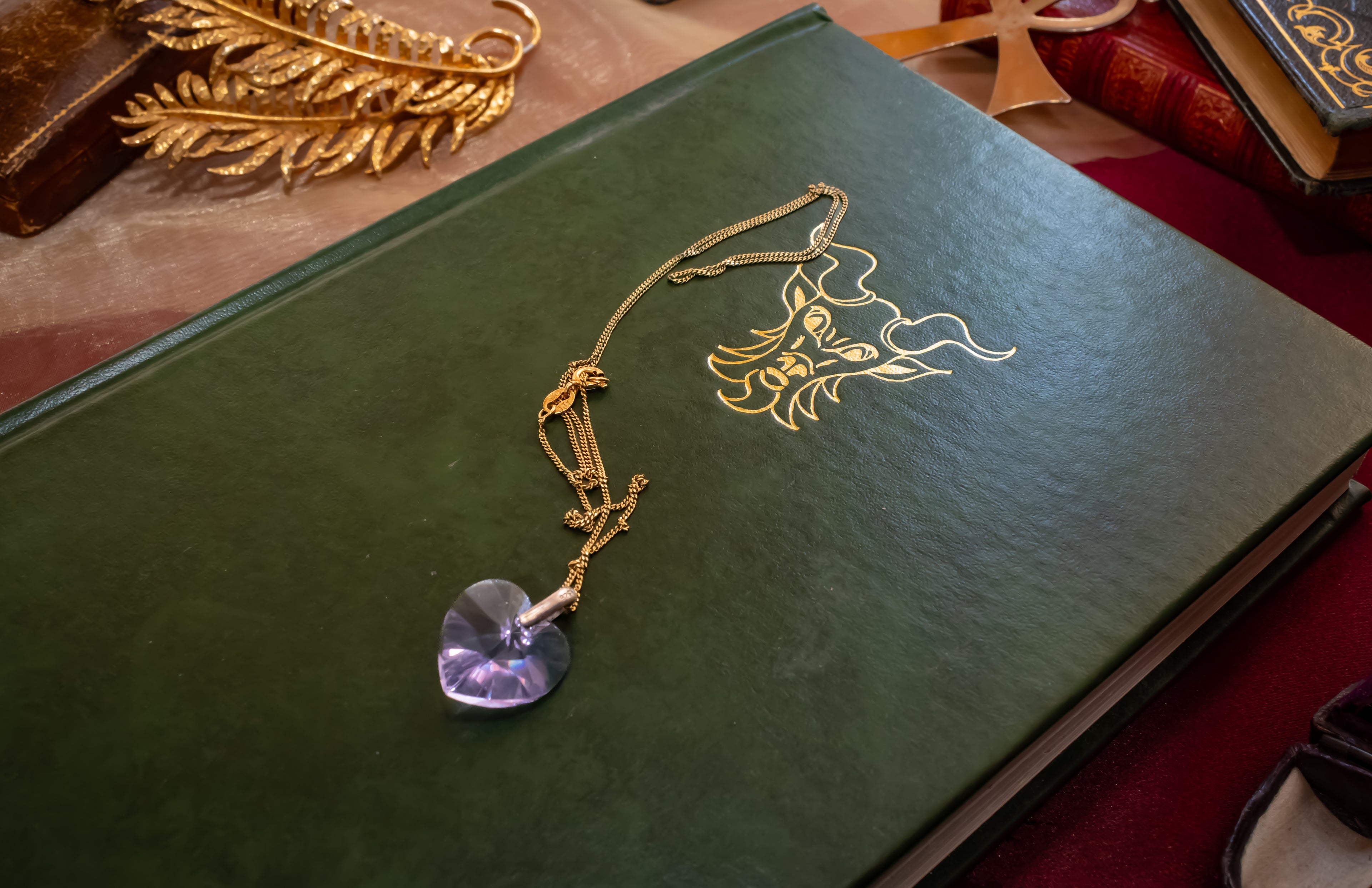 A gold chain necklace with a faceted purple crystal pendant resting on a dark green book with gold foil detailing.