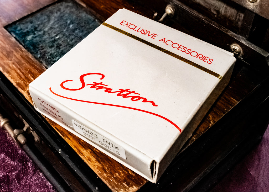A white Stratton exclusive accessories  pack box with red script lettering, resting on a dark wooden surface.