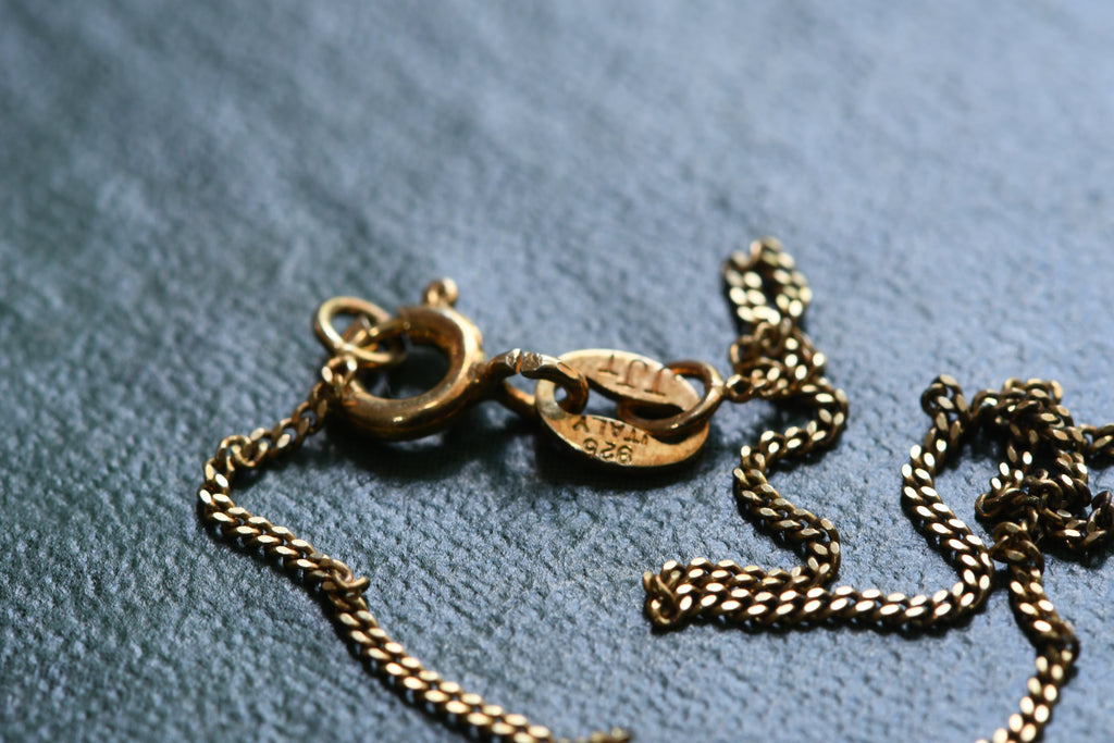 Close-up of a gold chain necklace clasp and tag on a textured blue-gray fabric background.