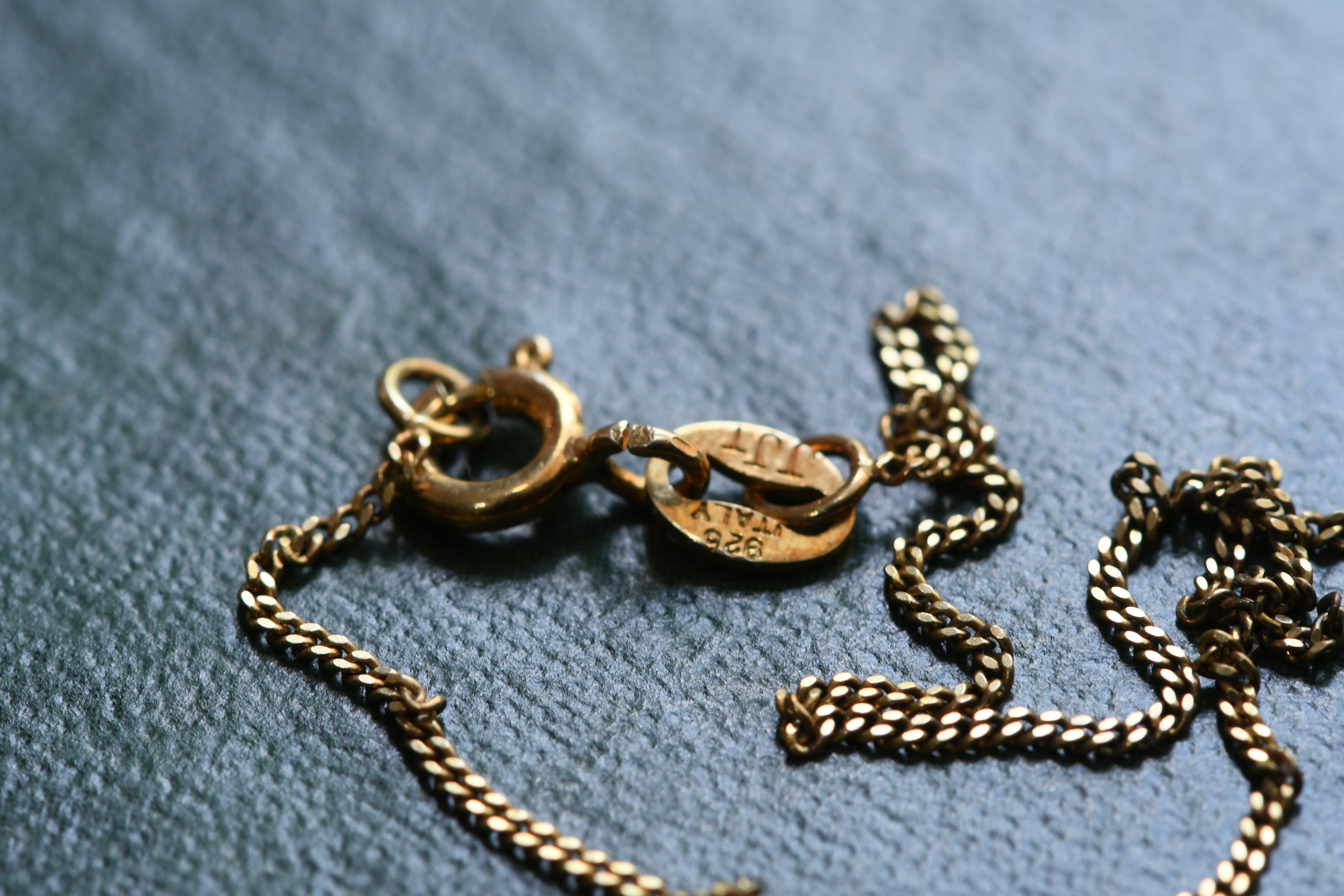 Close-up of a gold chain necklace clasp and tag on a textured blue-gray fabric background.