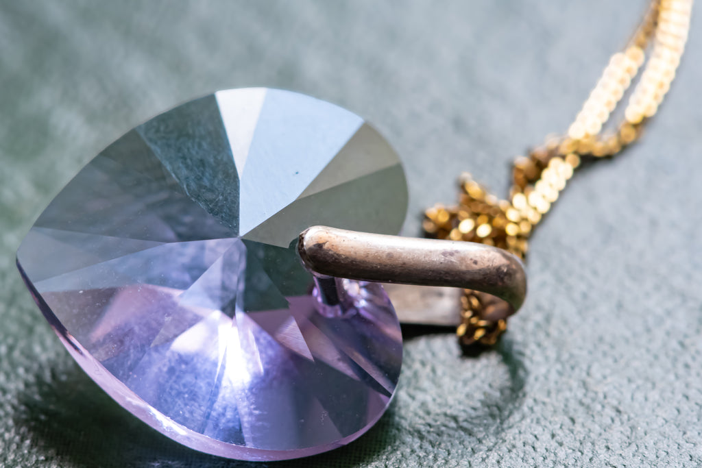 A close-up of a faceted heart-shaped purple crystal pendant on a gold chain, resting on a textured gray surface.