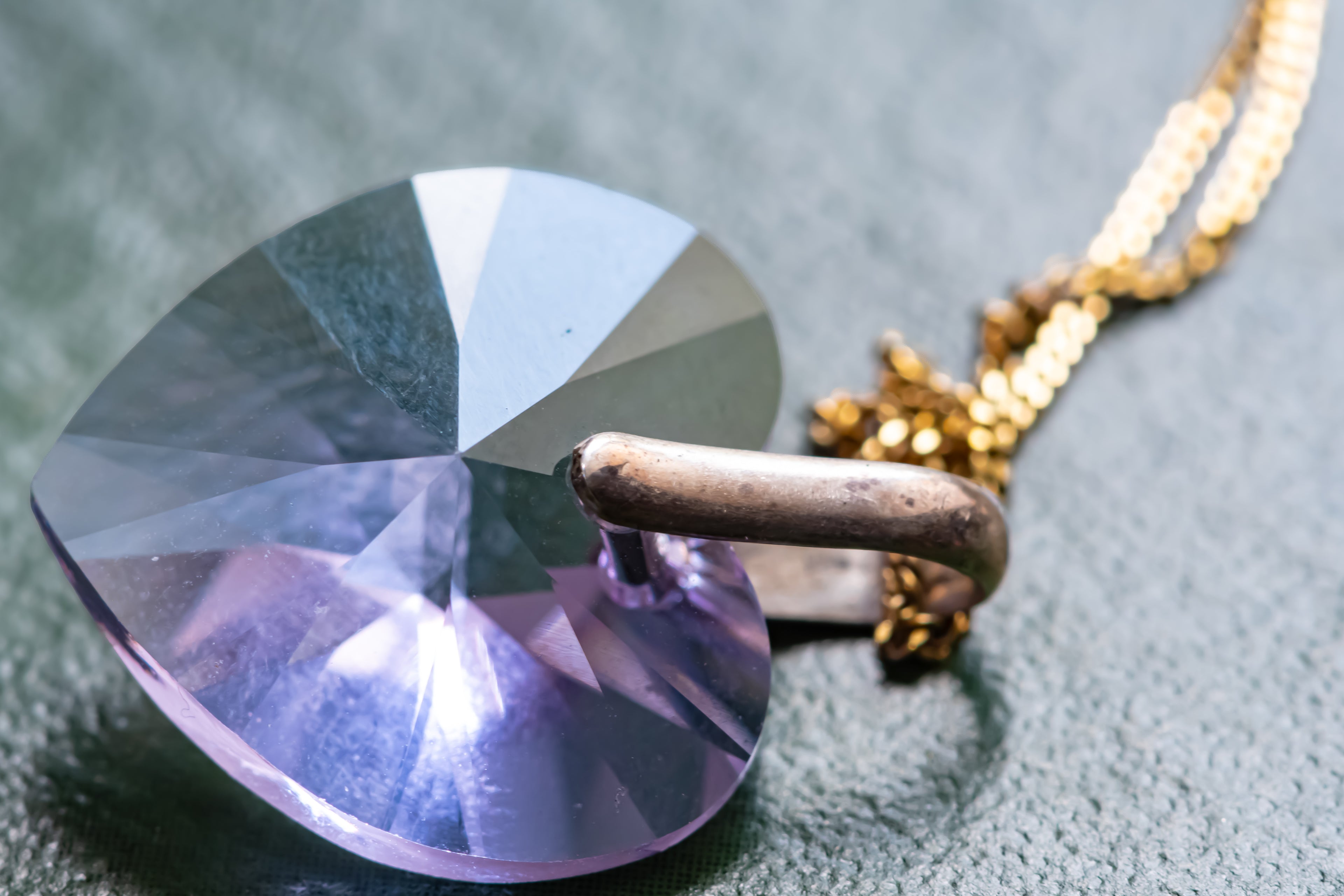 A close-up of a faceted heart-shaped purple crystal pendant on a gold chain, resting on a textured gray surface.