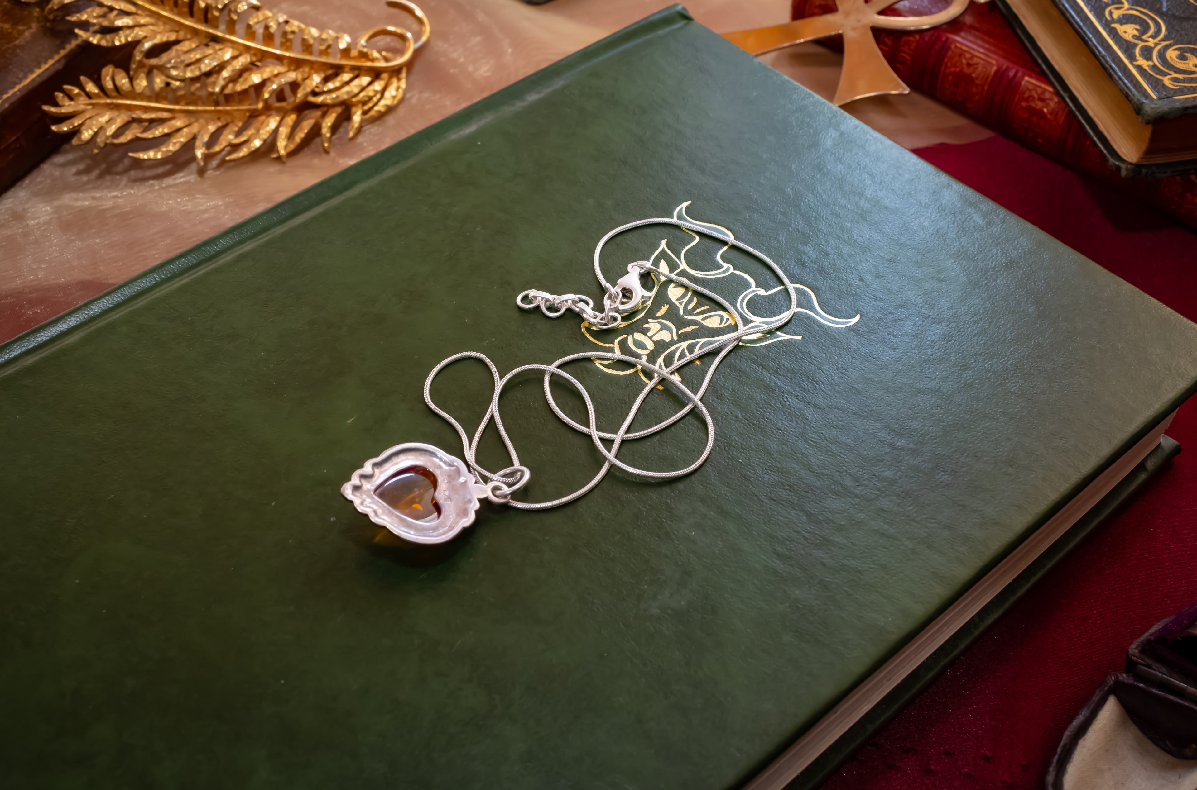 A silver-toned necklace with a heart-shaped pendant resting on a dark green book, surrounded by decorative gold and red items.