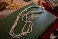A silver-toned multi-strand braided cord necklace draped over a dark green leather-bound book with gold detailing, set against a warm, vintage-themed background with books and a feather ornament.