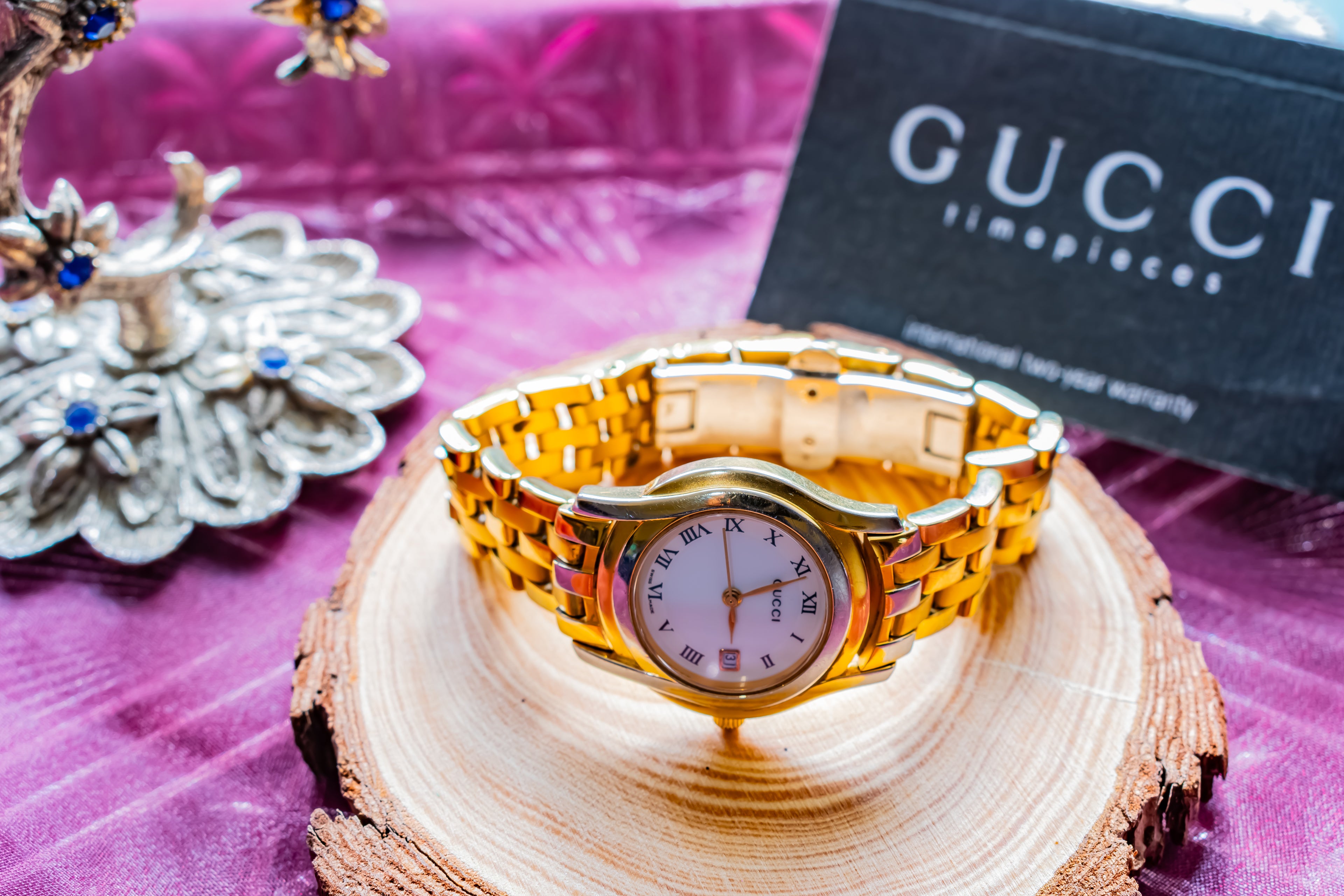 A gold-tone Gucci watch with a two-tone gold and silver link bracelet, displayed on a wooden slice coaster. A black Gucci Simplices warranty card is visible in the background on a purple textured surface.