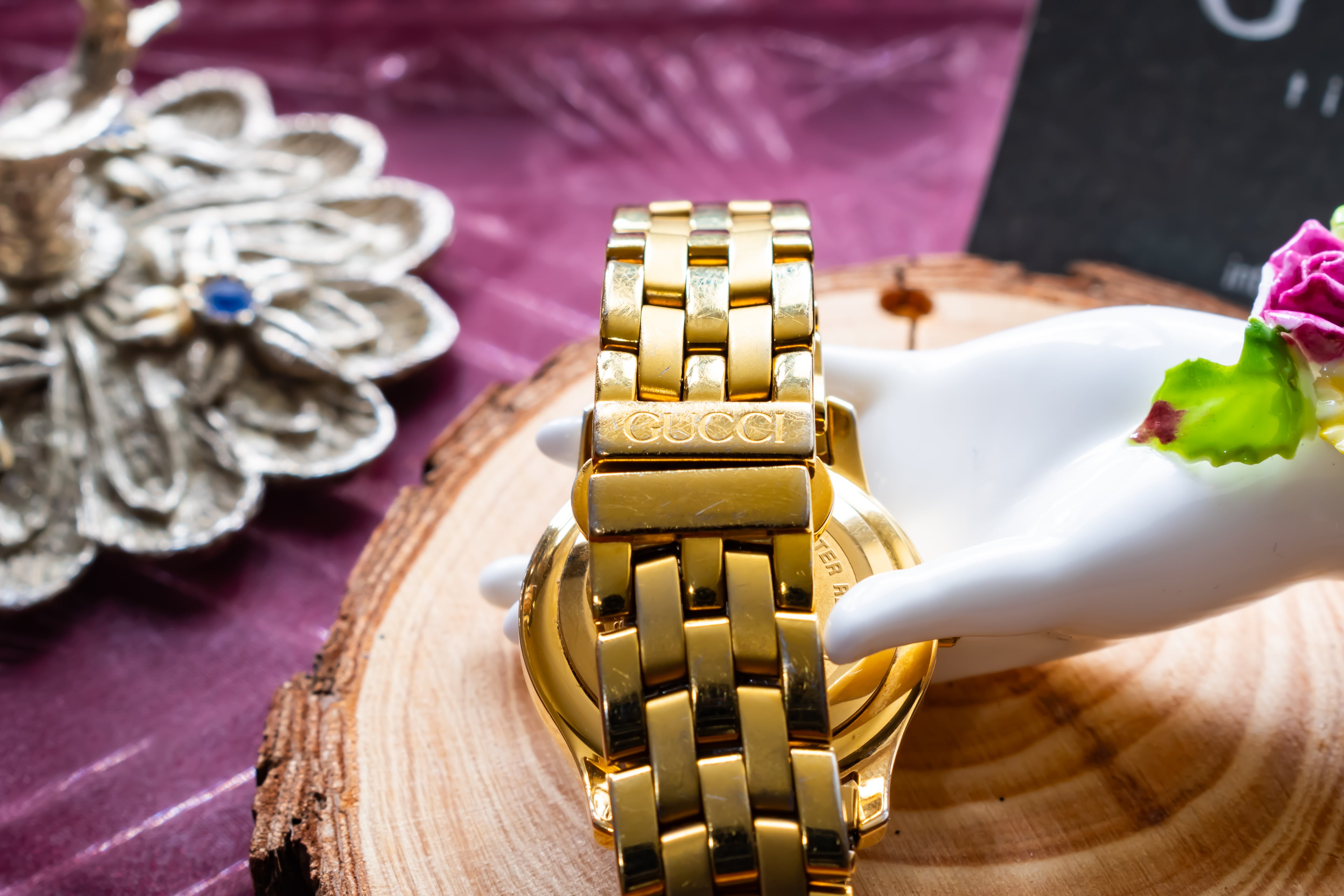 A gold-tone Gucci metal link watch band shown from the back, resting on a white ceramic hand display over a wood slice, with a purple fabric background and decorative props.