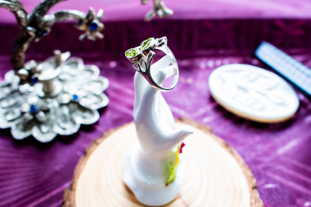 A silver ring with a green crystal cluster setting, displayed on a white ceramic bird figurine on a wooden slice coaster, set against a purple fabric background.