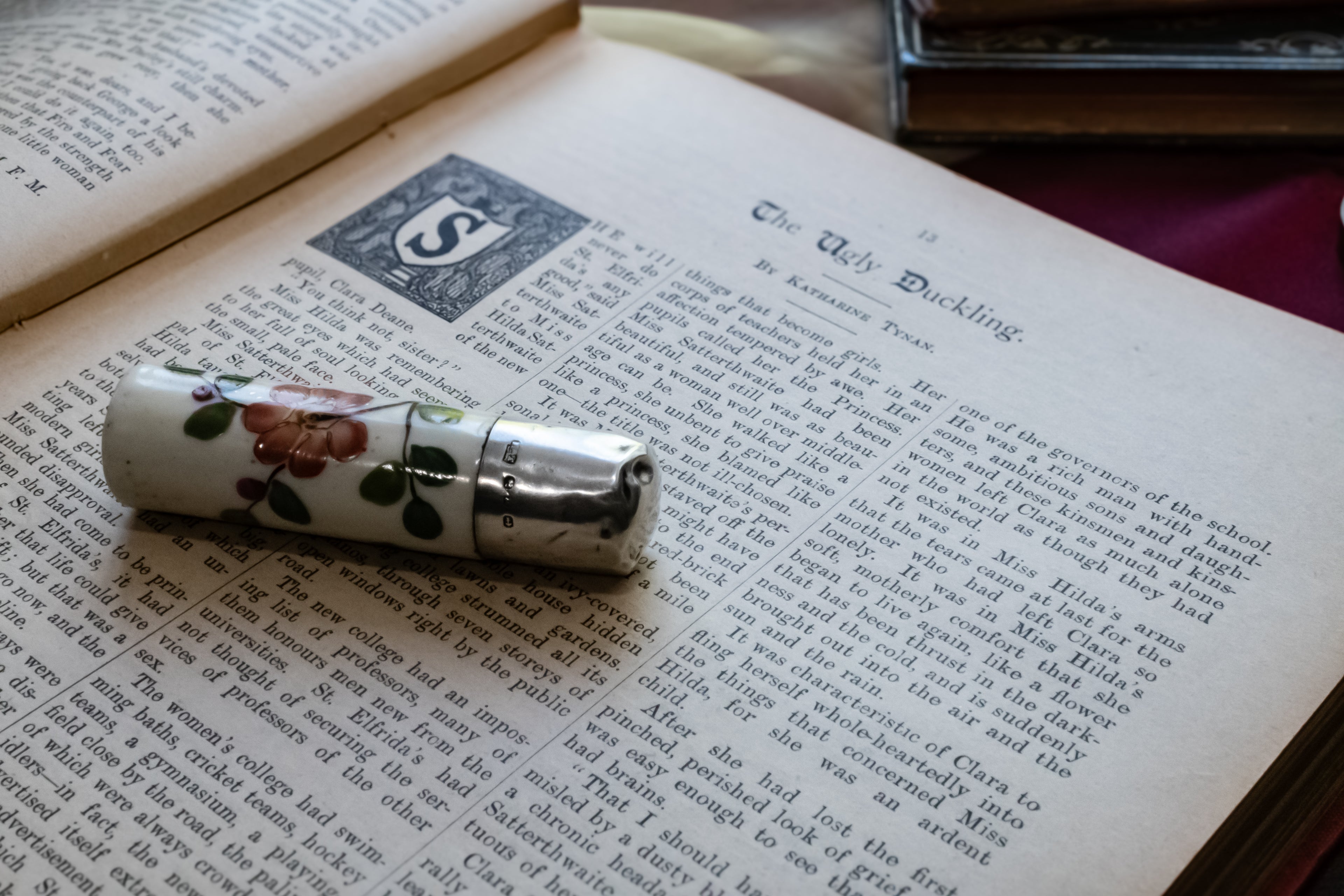 A vintage silver and white perfume bottle with floral detailing rests on an open antique book.
