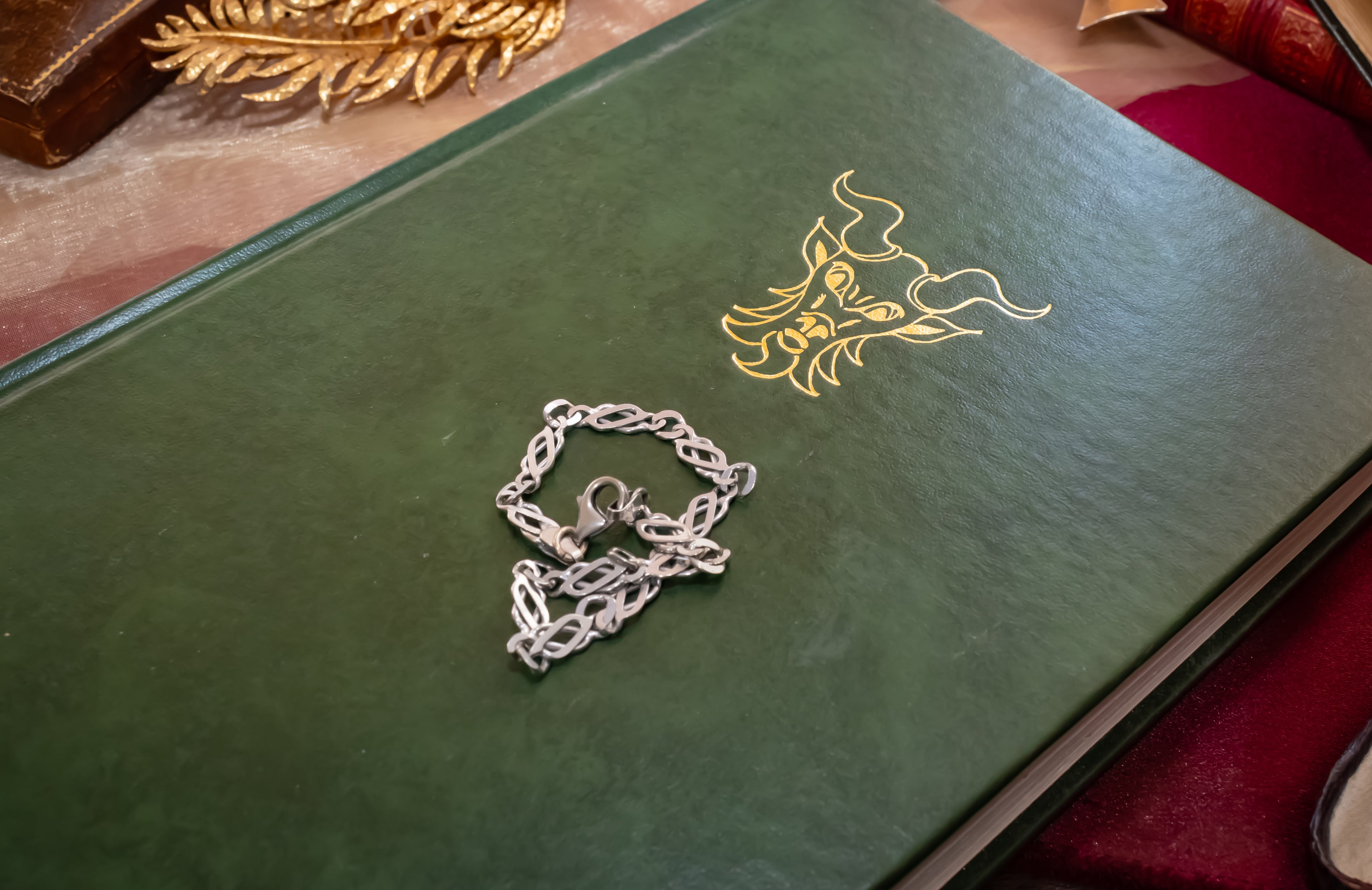 A silver chain bracelet with a lobster clasp and decorative pendant, resting on a dark green book with a gold dragon illustration.