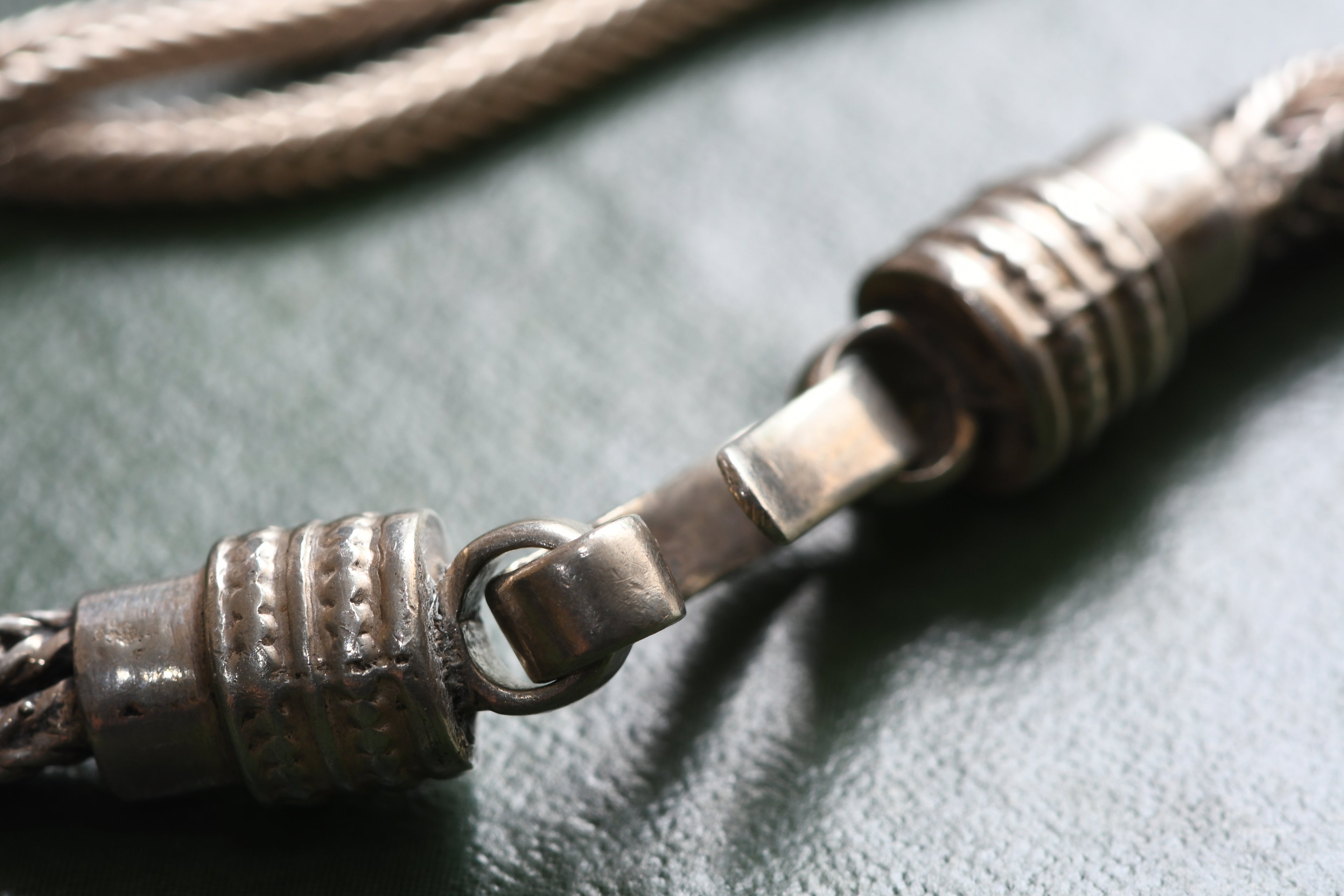 A close-up of a silver-toned metal chain with textured cylindrical links and a ring clasp, resting on a dark green textured surface.