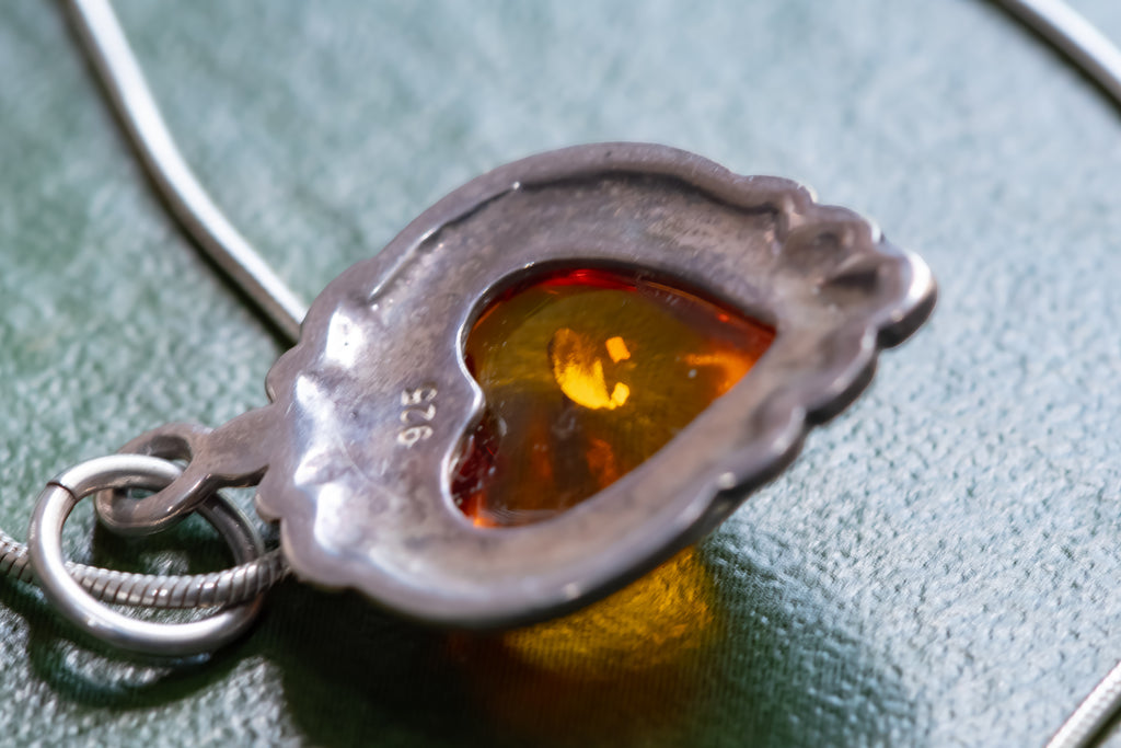 A close-up of a silver pendant necklace featuring a heart-shaped amber cabochon, resting on a textured dark green surface.