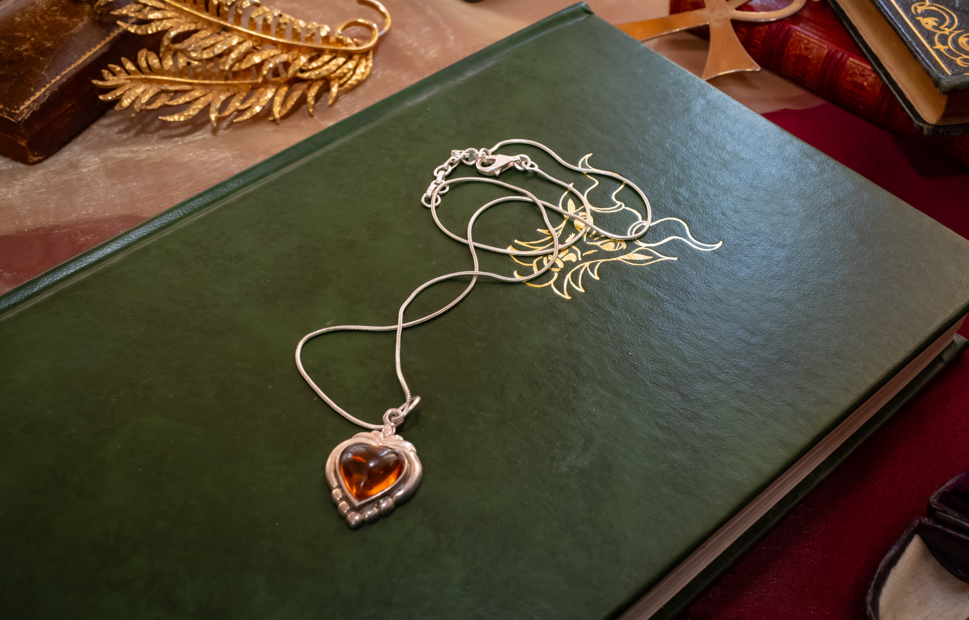 A silver heart-shaped pendant necklace with an amber-orange heart inlay, displayed on a dark green book with gold foil artwork.
