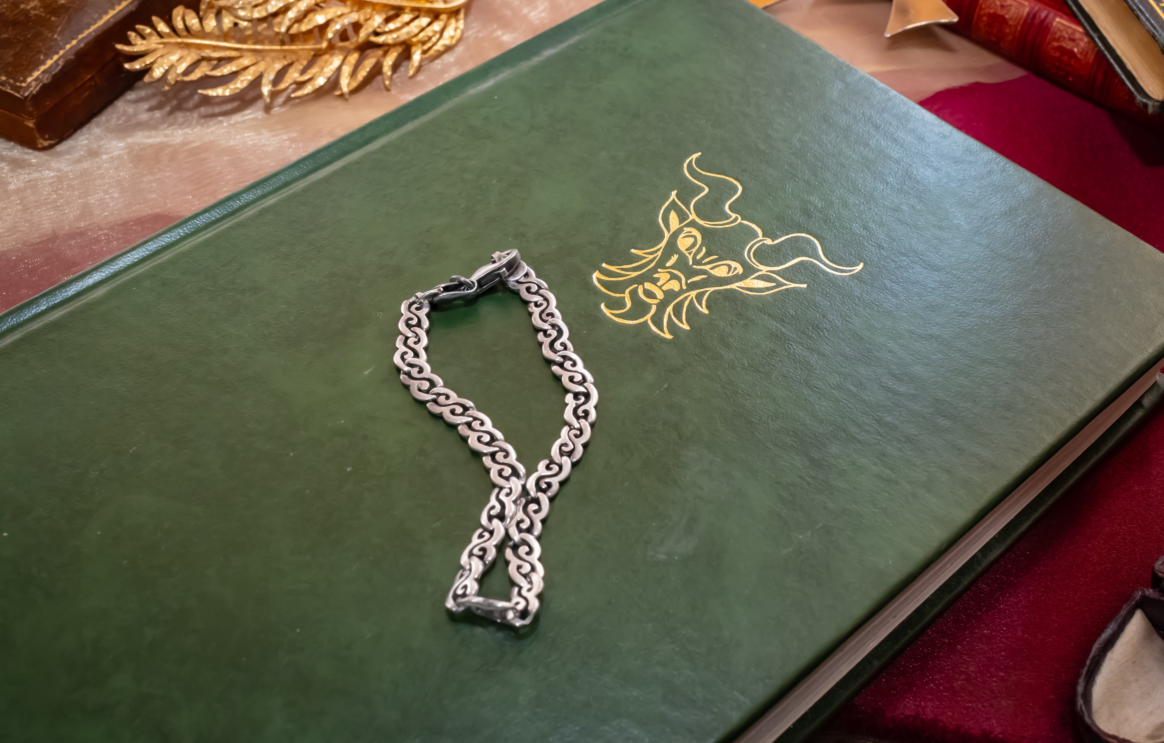 A silver chain accessory with a lobster clasp rests on a dark green book with a gold dragon illustration. The book is placed on a red fabric surface with other decorative books in the background.