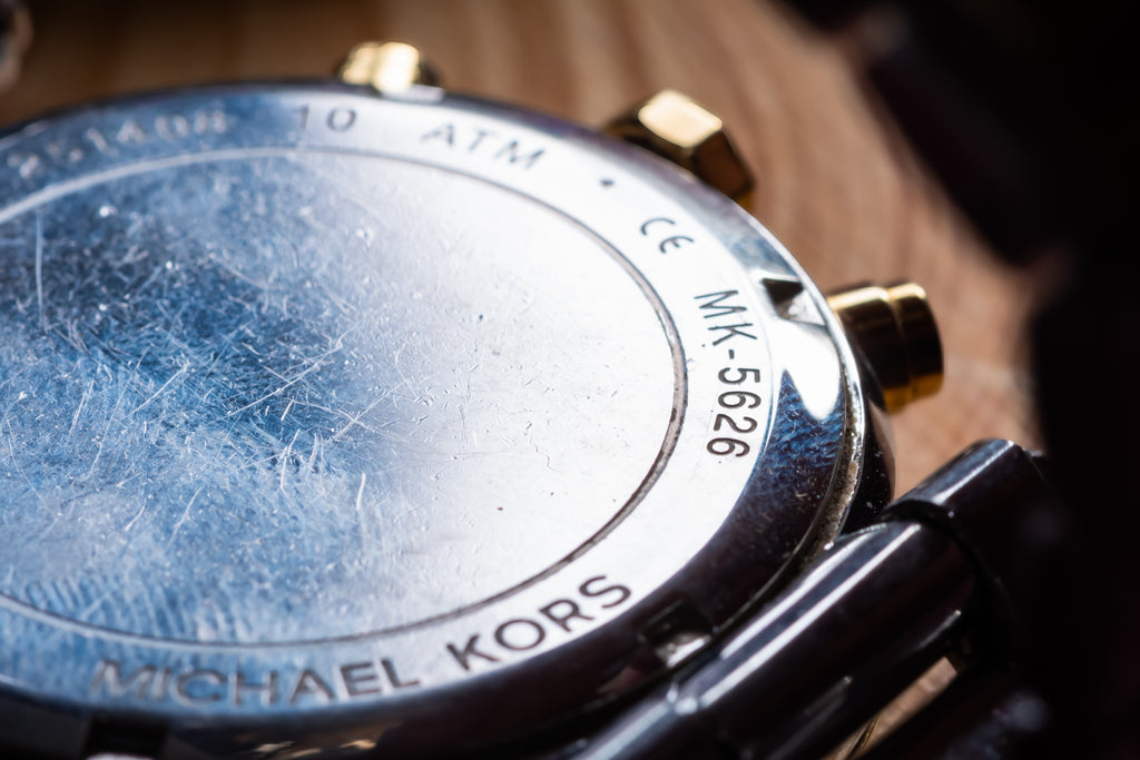 Close-up of the back case of a Michael Kors watch, showing a scratched silver-tone metal surface with gold-tone pushers and a dark strap on a wooden surface.