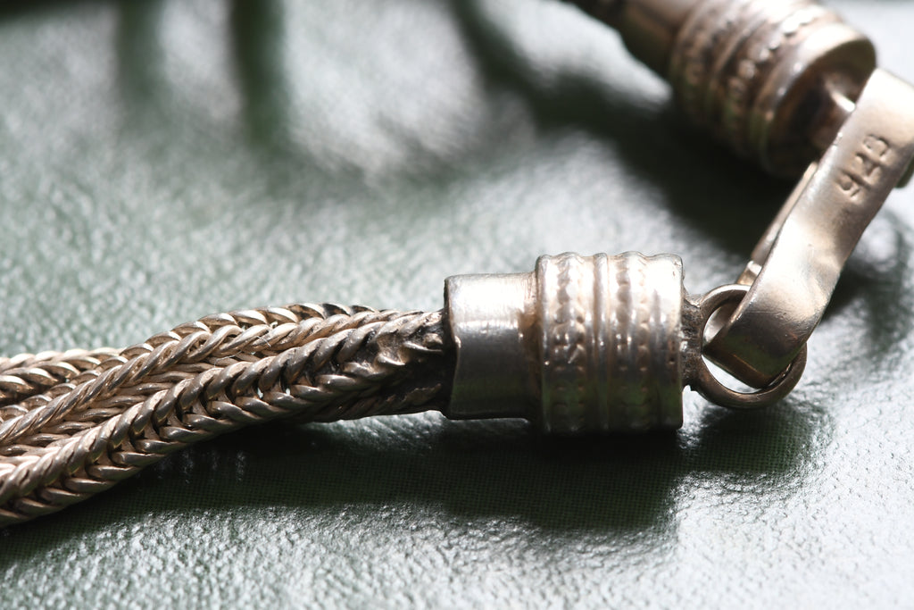 Close-up of a braided metal chain with a textured cylindrical clasp and a lobster clasp on a dark green textured surface.