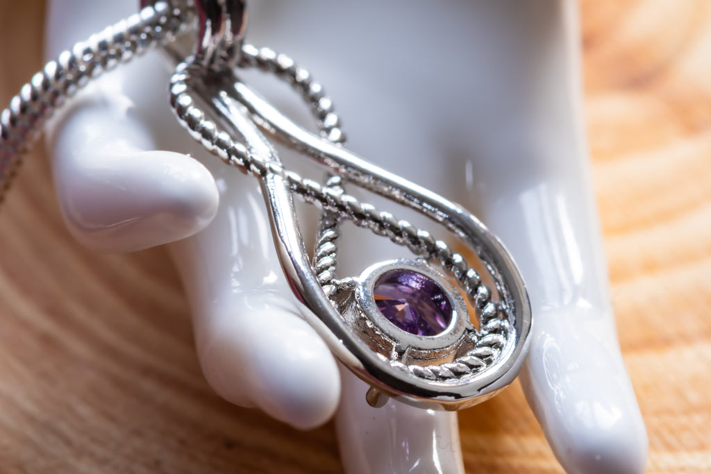 A silver teardrop pendant necklace with a central oval purple gemstone, displayed on a glossy white ceramic mannequin hand over a wooden surface.