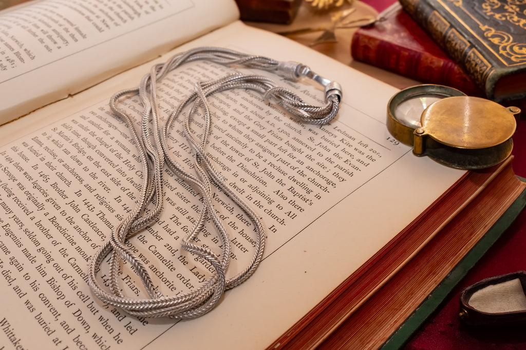 A silver-toned metal chain necklace with a textured braid pattern and a hook clasp, draped over the pages of an open vintage book. The scene is styled with antique books, a brass pocket watch, and a red velvet surface in the background.