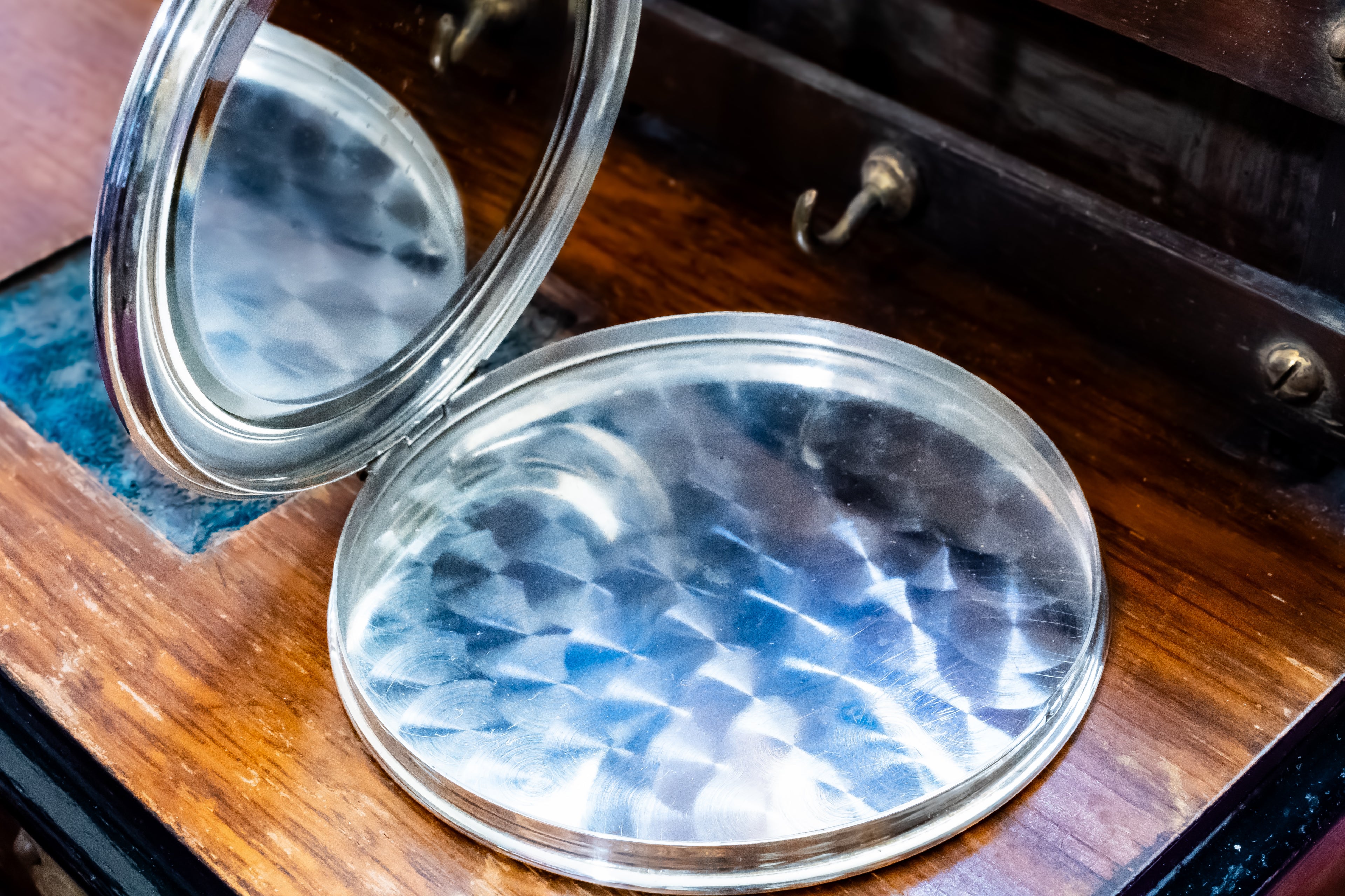 A round silver-toned compact mirror with a hammered glass finish, shown open on a dark wooden surface.