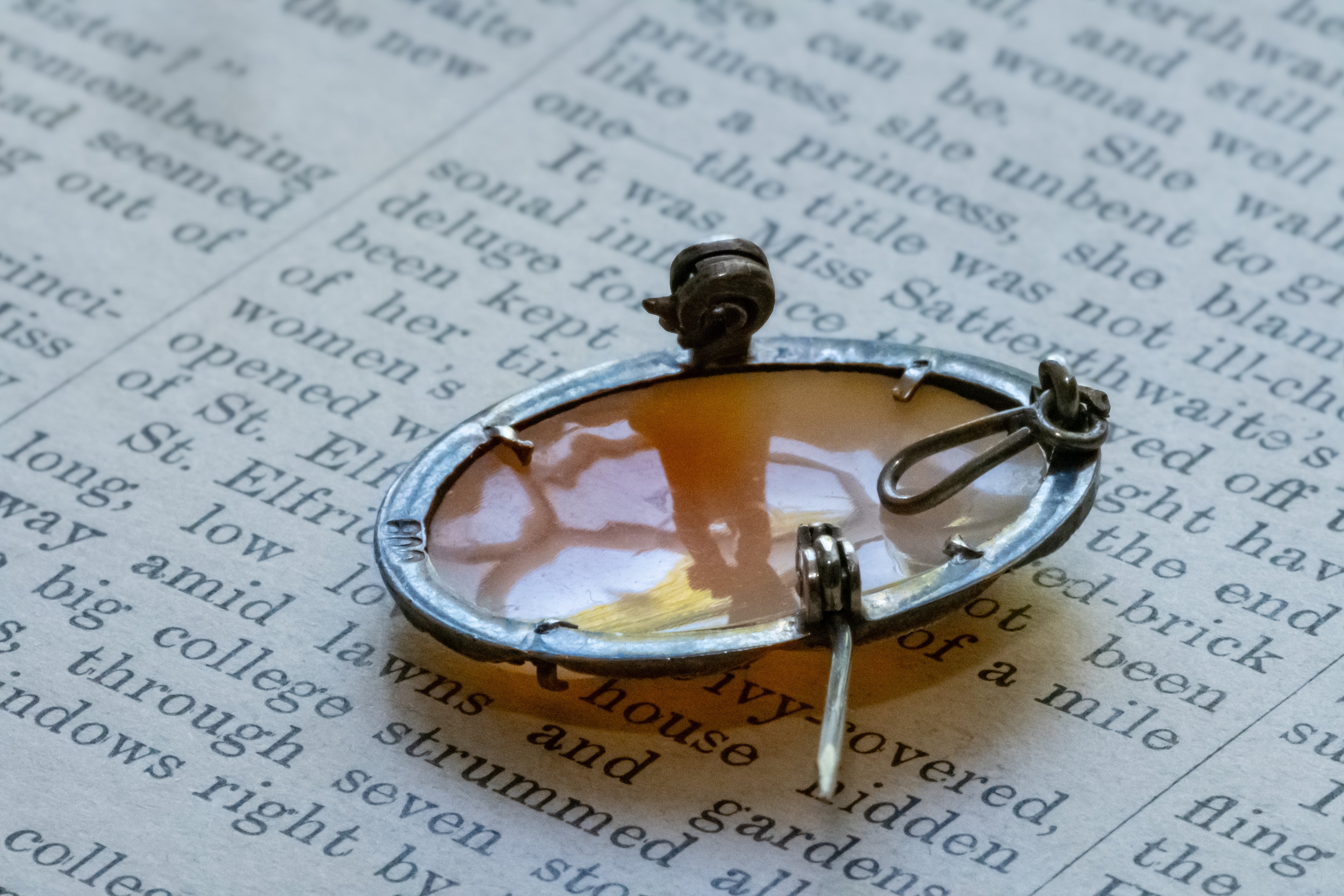 A silver-toned oval pin with a translucent amber-orange center and a small dark metal top, photographed close up on printed book text.