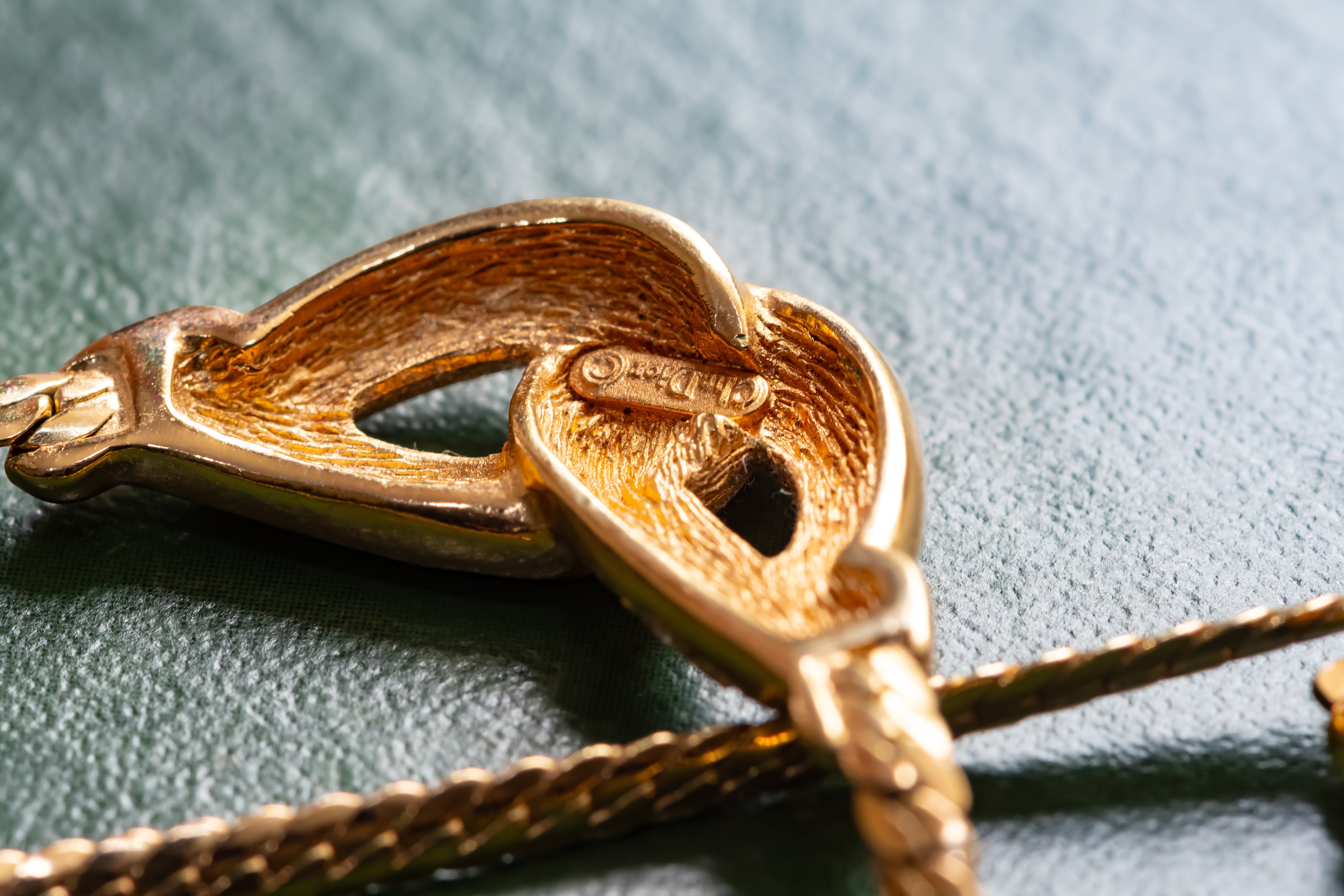 Close-up of a gold heart-shaped pendant clasp on a gold chain, resting on a textured dark green surface.