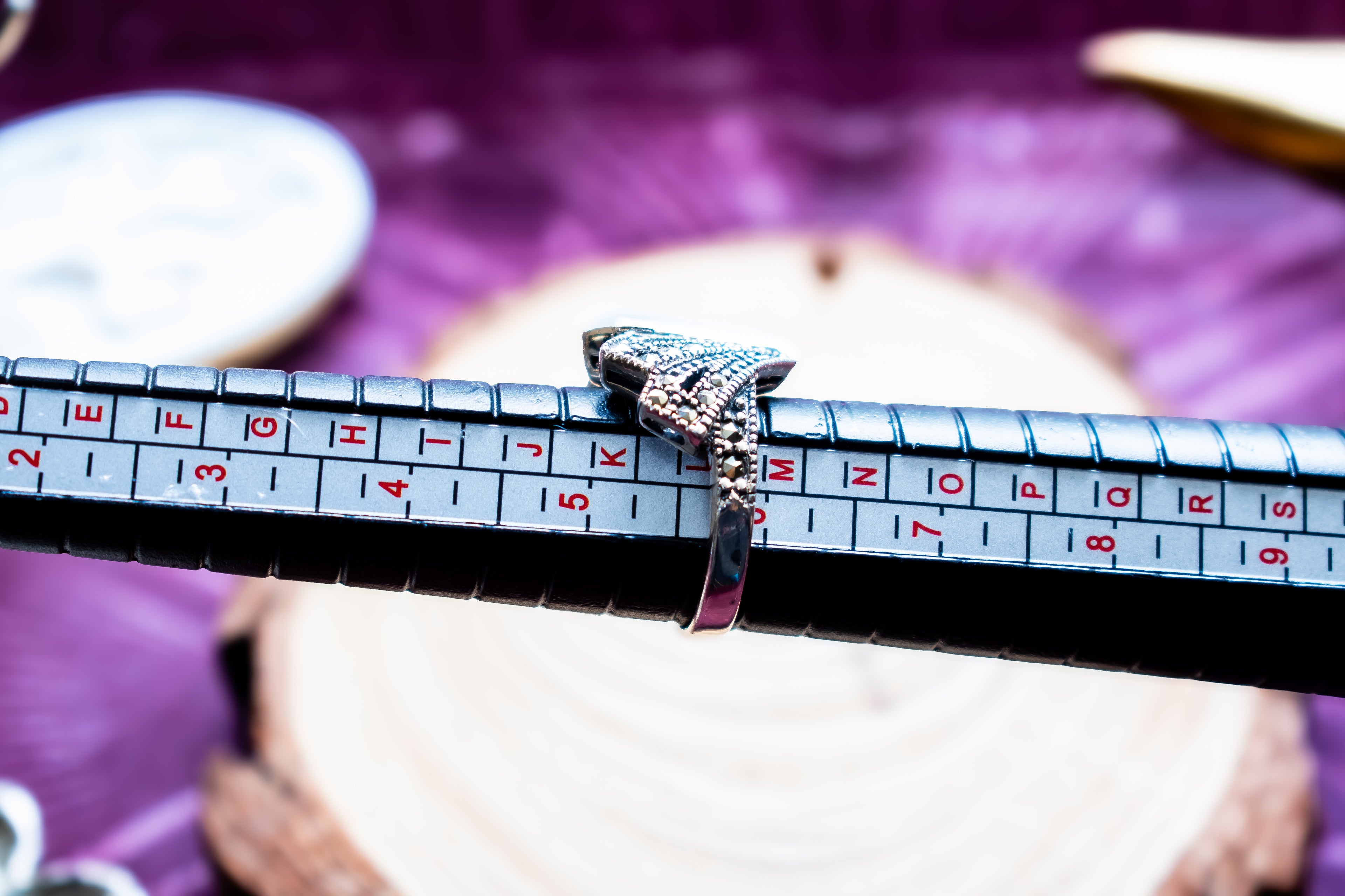 A silver-toned ring with a textured, diamond-like top is displayed on a black ring sizer measuring tool, set against a purple fabric background with a blurred wooden slice.