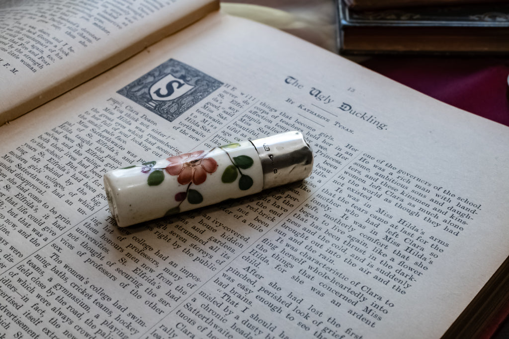 A vintage ceramic scent bottle with a floral pattern and silver-toned metal casing, resting on an open antique book.