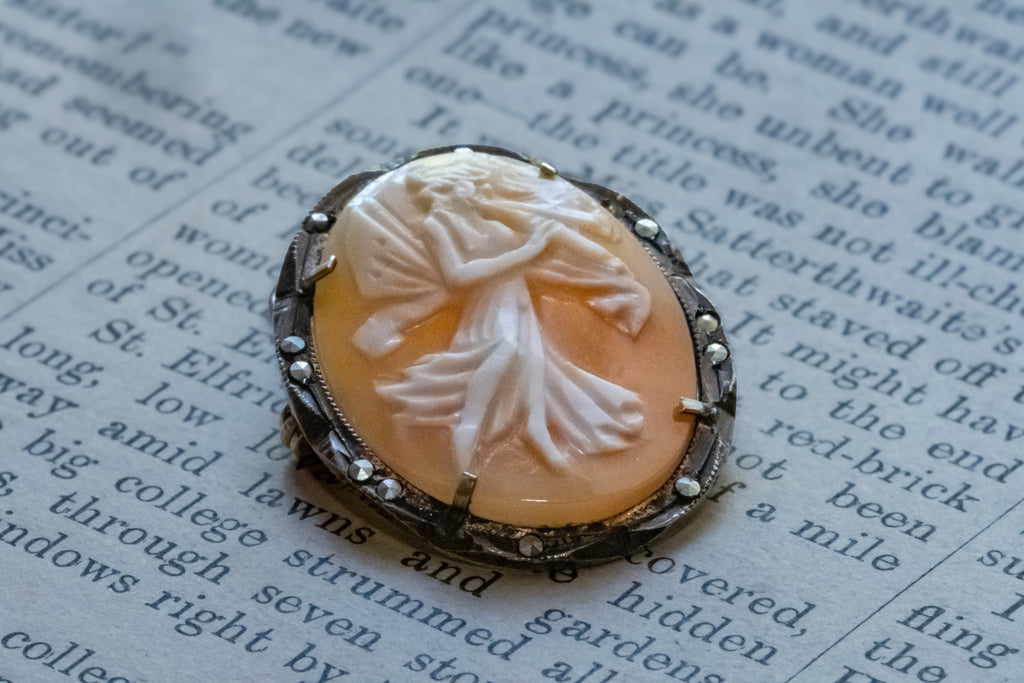 A vintage-style carved shell  pendant with a white fairy motif set in a silver-toned bezel, resting on printed book pages.