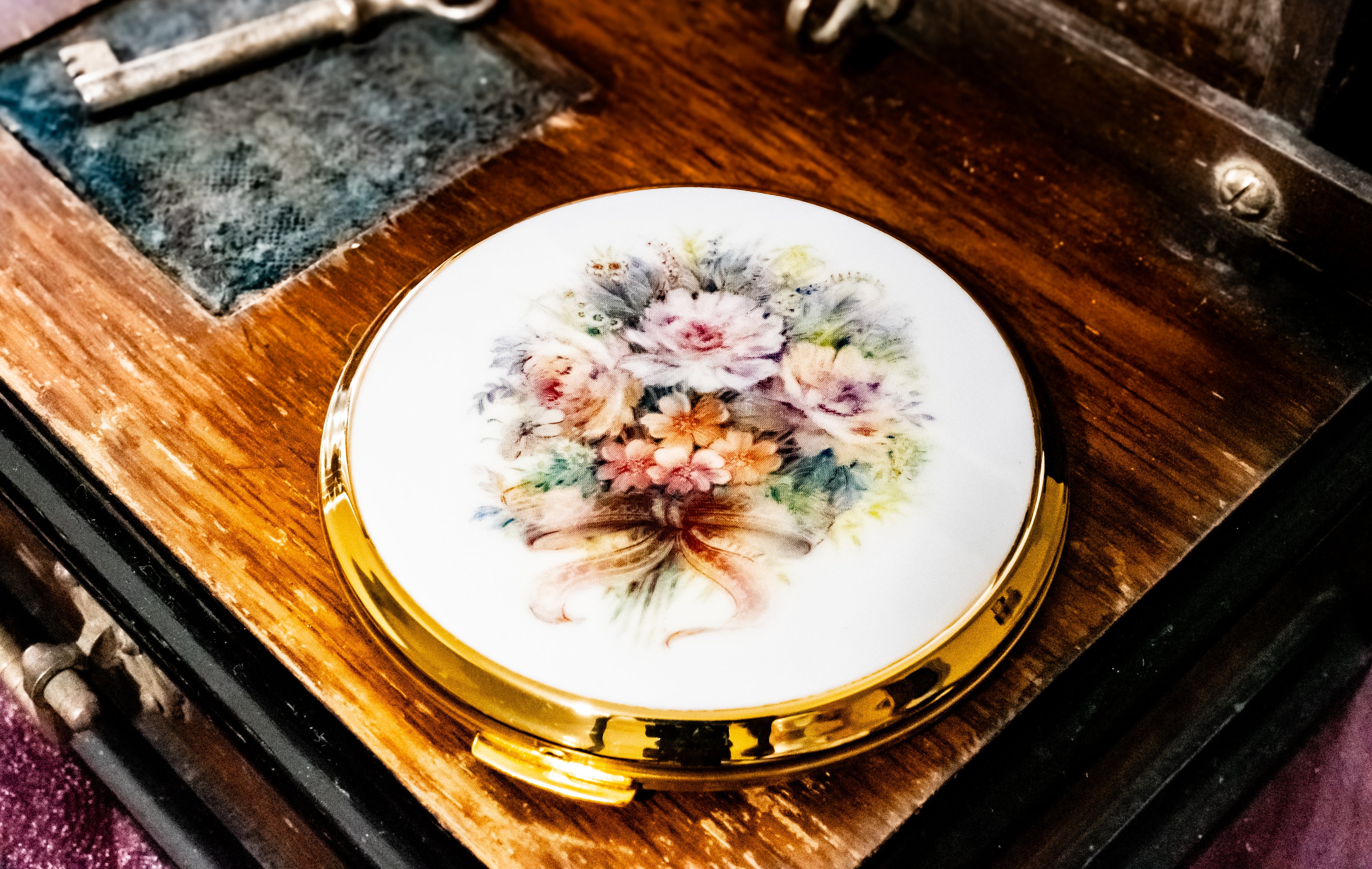 A round compact mirror with a white floral face and a shiny gold rim, resting on a vintage wooden box with a key nearby.