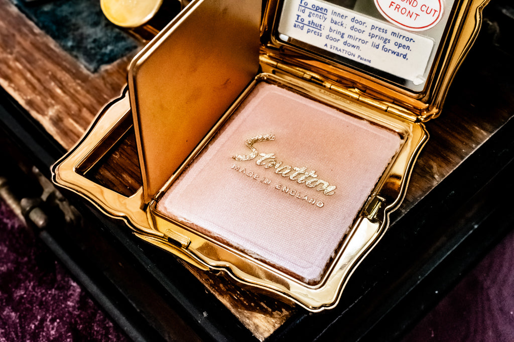 An open gold compact containing a shimmering light pink pressed powder, with the brand name 'Stratton' embossed on the powder puff. The compact is shown resting on a dark wooden surface.