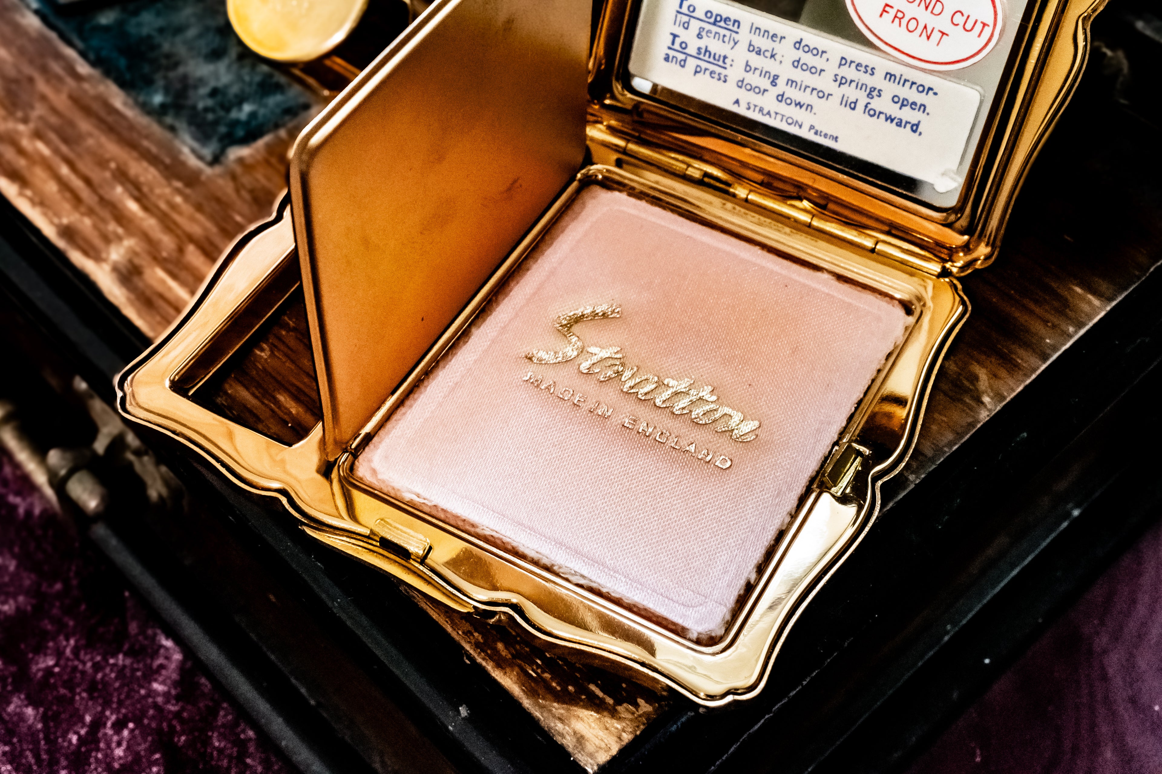 An open gold compact containing a shimmering light pink pressed powder, with the brand name 'Stratton' embossed on the powder puff. The compact is shown resting on a dark wooden surface.