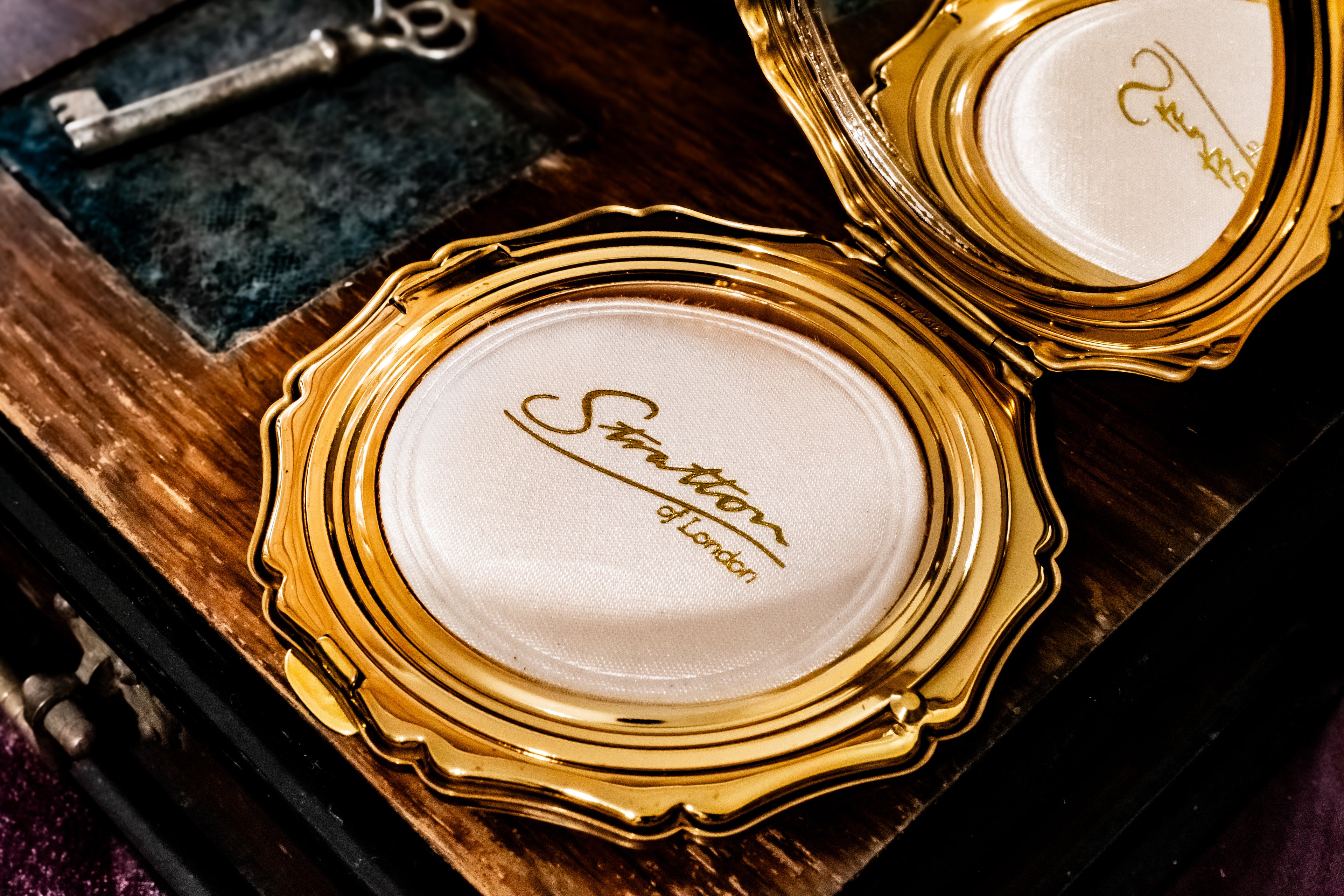A close-up of a gold oval compact mirror with a white pressed powder pan and the brand name 'Stratton of London' printed in gold on the lid, resting on a dark wooden surface next to a vintage key.