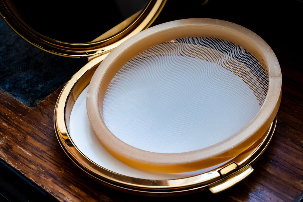 An open round compact with a gold-tone metal frame and mirror, containing a circular beige ring-shaped item with a fine mesh screen over a white base, photographed on a dark wooden surface.