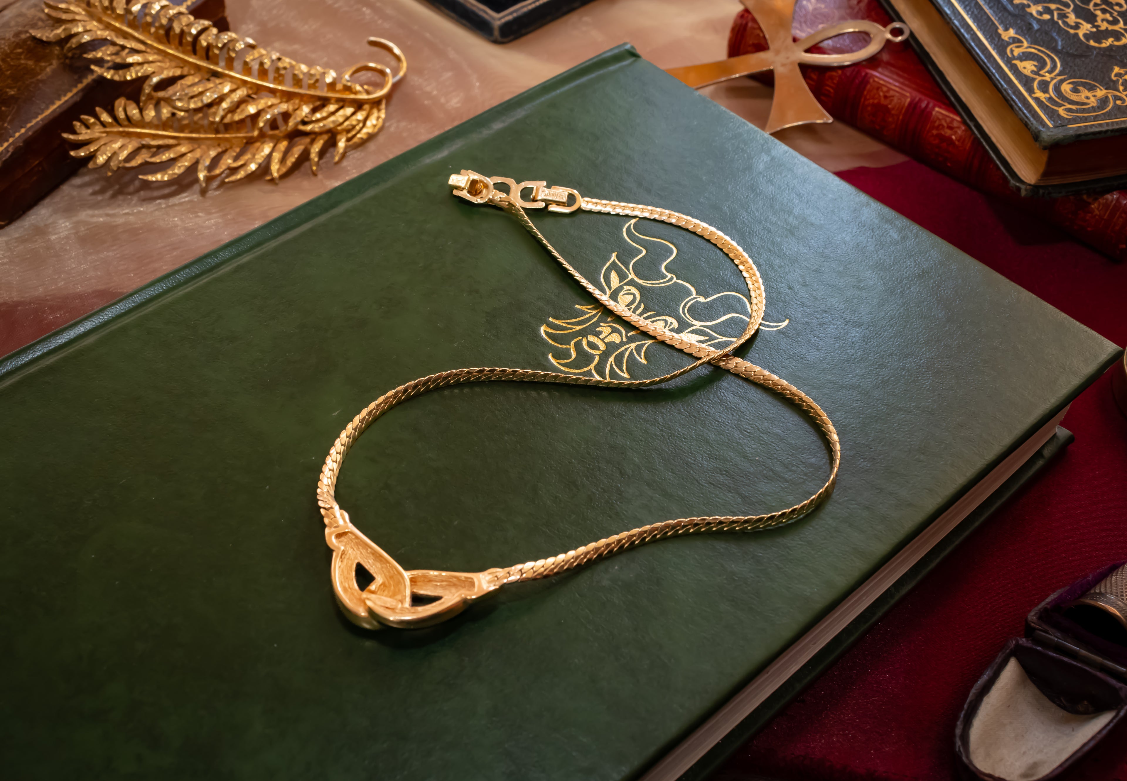 A gold chain necklace with a decorative pendant, draped elegantly over a dark green leather-bound book on a red velvet surface.