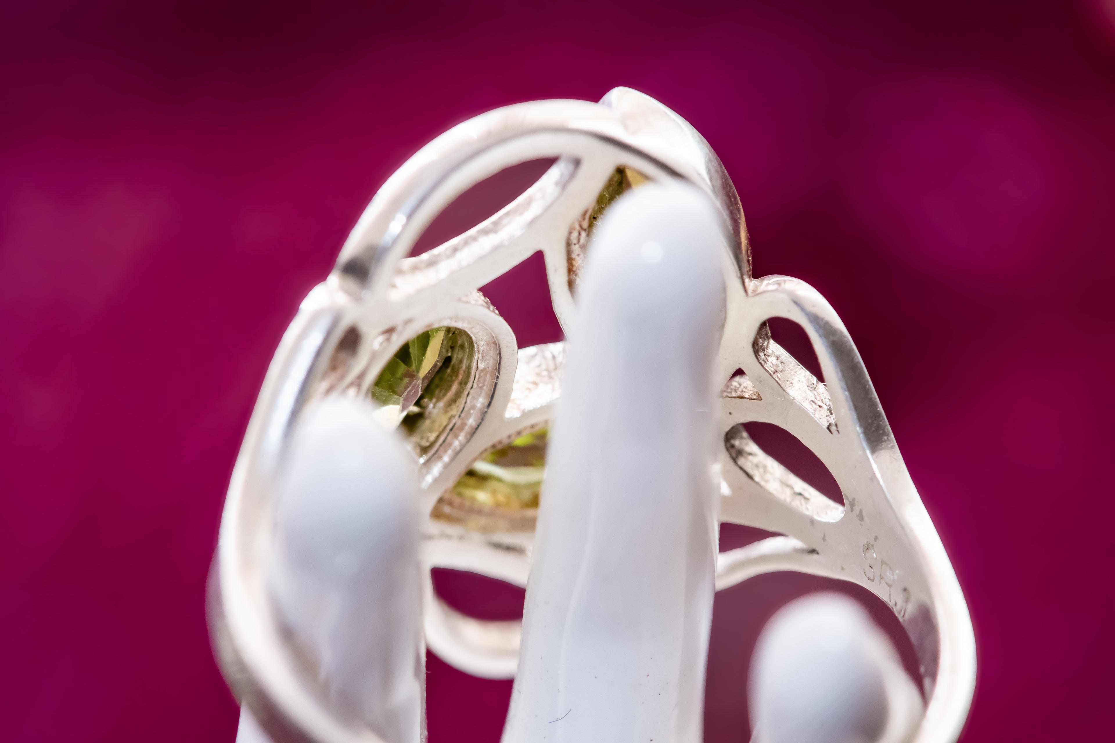 A close-up of a silver filigree ring with a green gemstone, displayed on a white ring stand against a vibrant magenta background.