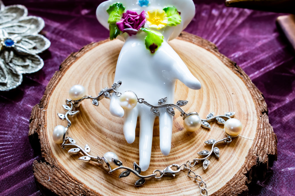 A silver vine-and-pearl necklace displayed on a white ceramic hand-shaped stand, set on a round wood slice over a purple fabric background.