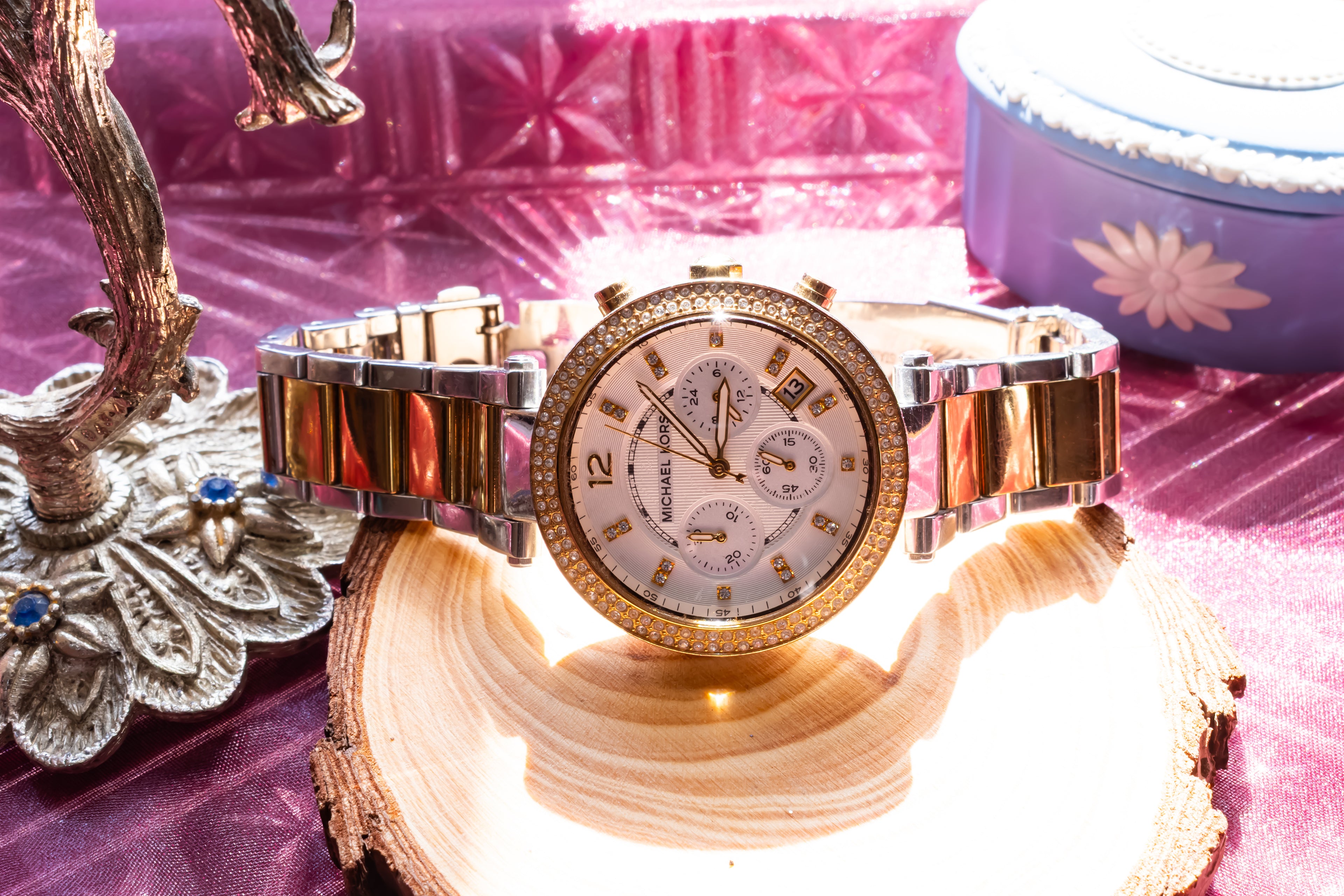 A luxury chronograph watch with a gold and silver two-tone metal bracelet, a gold bezel, and a white dial with sub-dials, displayed on a wood slice with pink and purple fabric background.