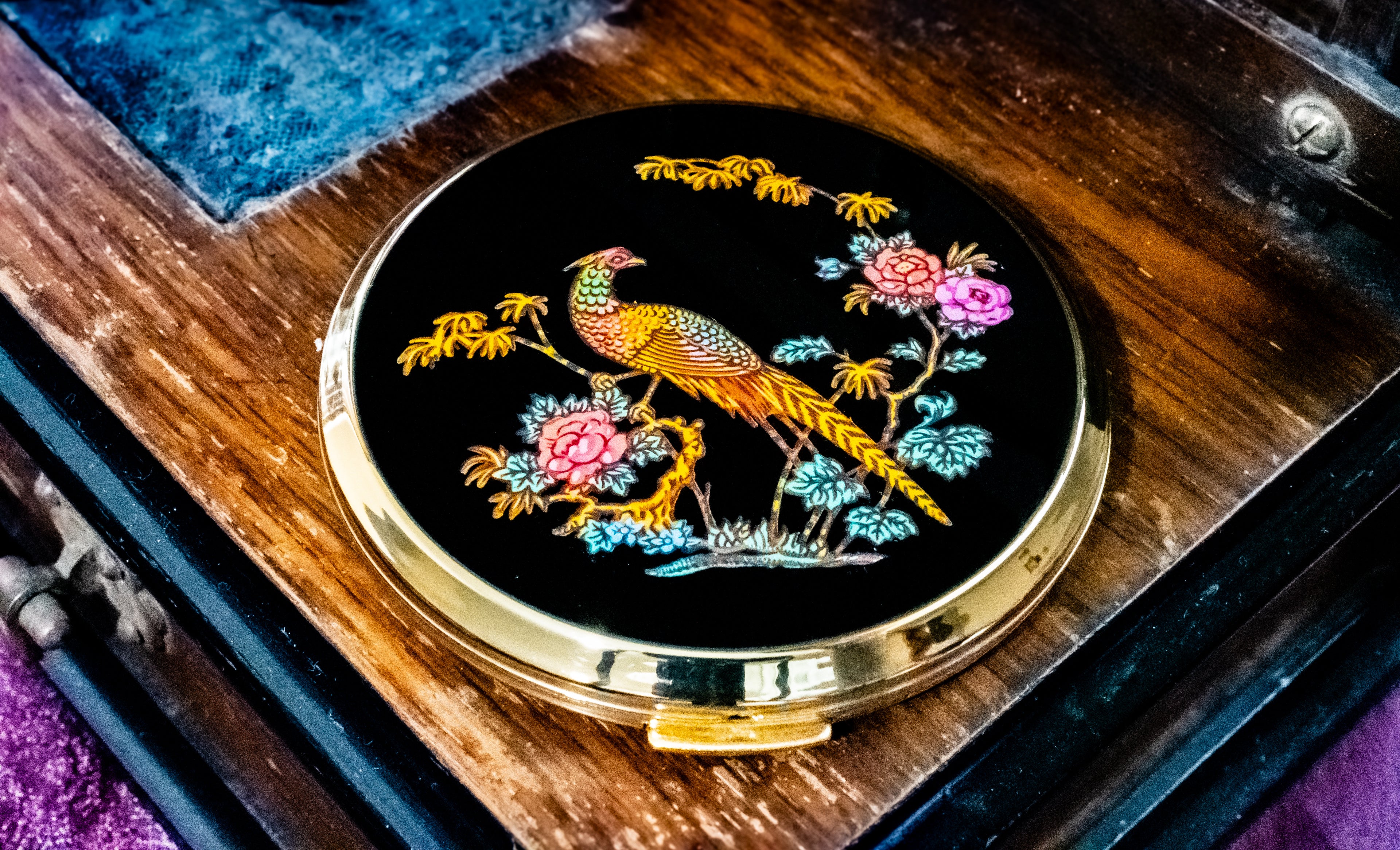 A round black compact mirror with a gold-tone metal rim, featuring an intricate embroidered peacock and floral design in gold, pink, and blue on a dark background, resting on a wooden surface.