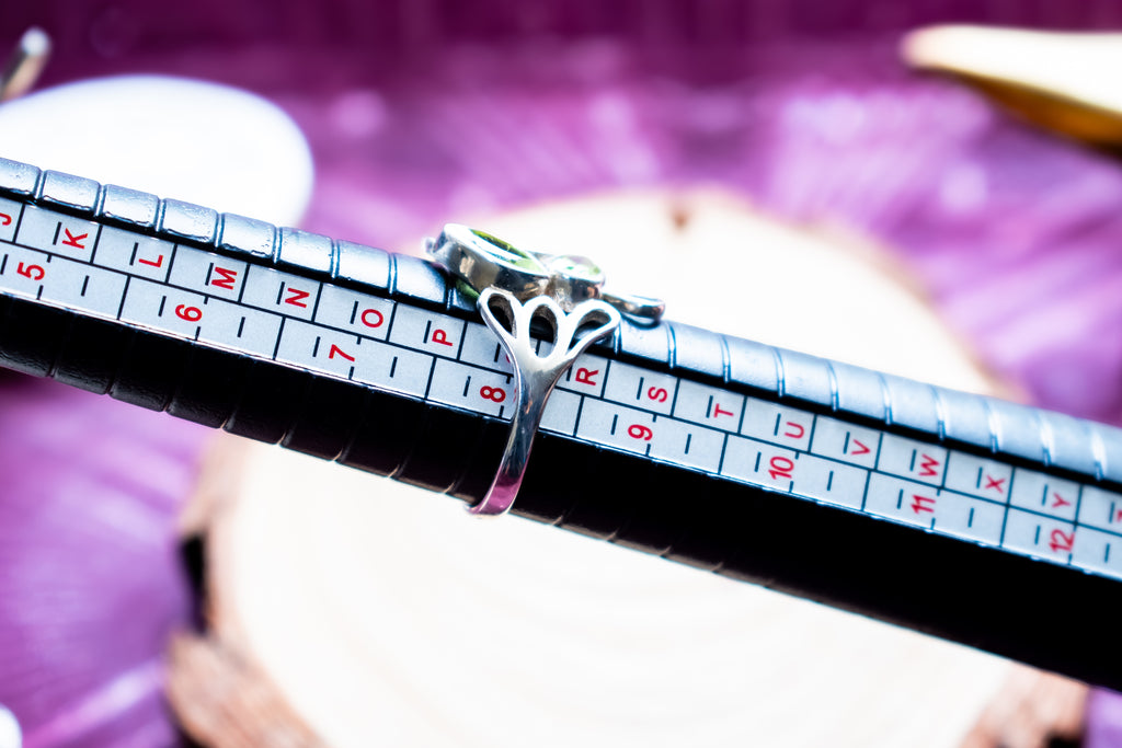 A silver ring with a butterfly-shaped band and a small green stone is shown on a black ring sizer tool with size markings, set against a blurred purple background.