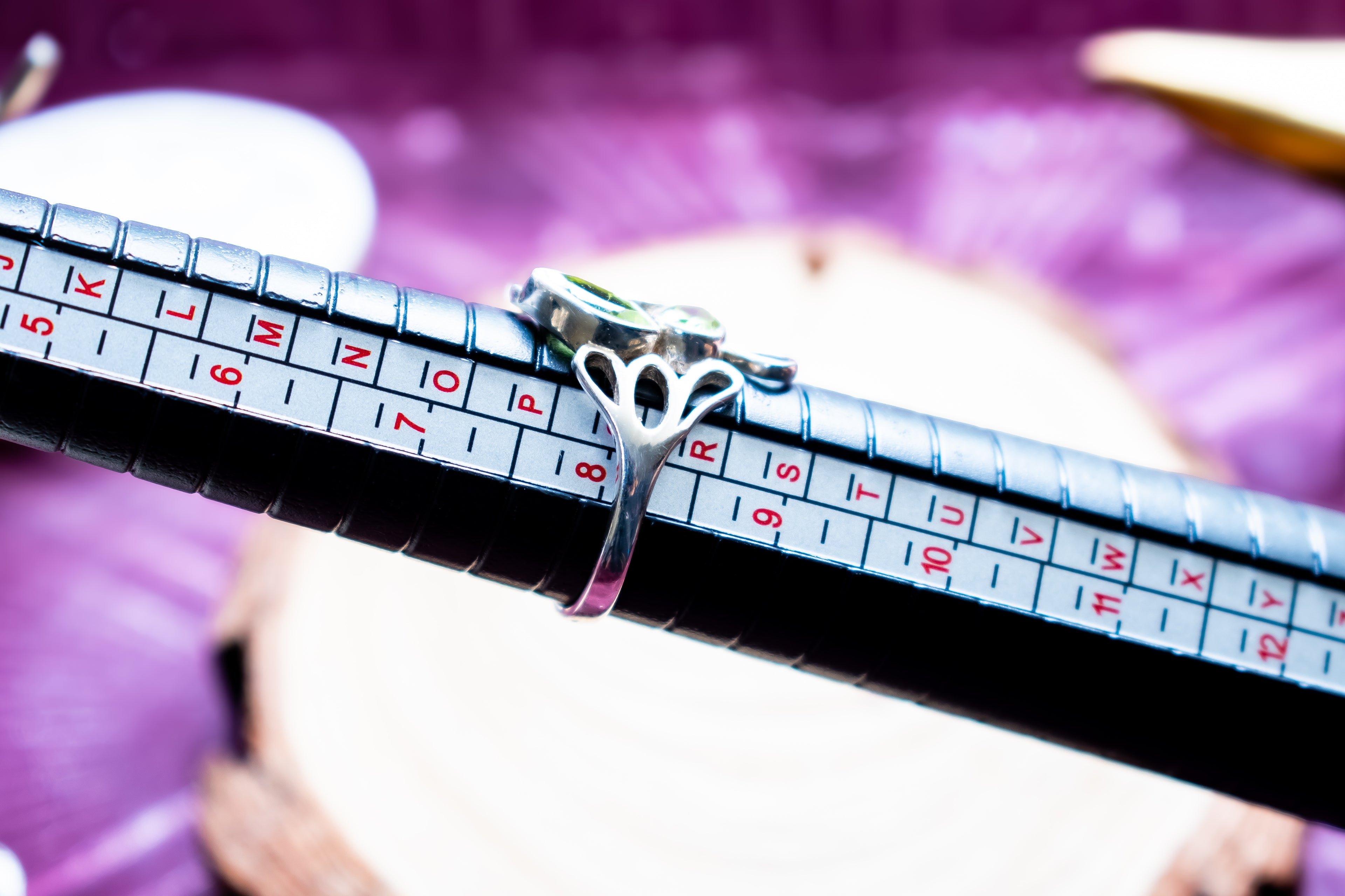 A silver ring with a butterfly-shaped band and a small green stone is shown on a black ring sizer tool with size markings, set against a blurred purple background.