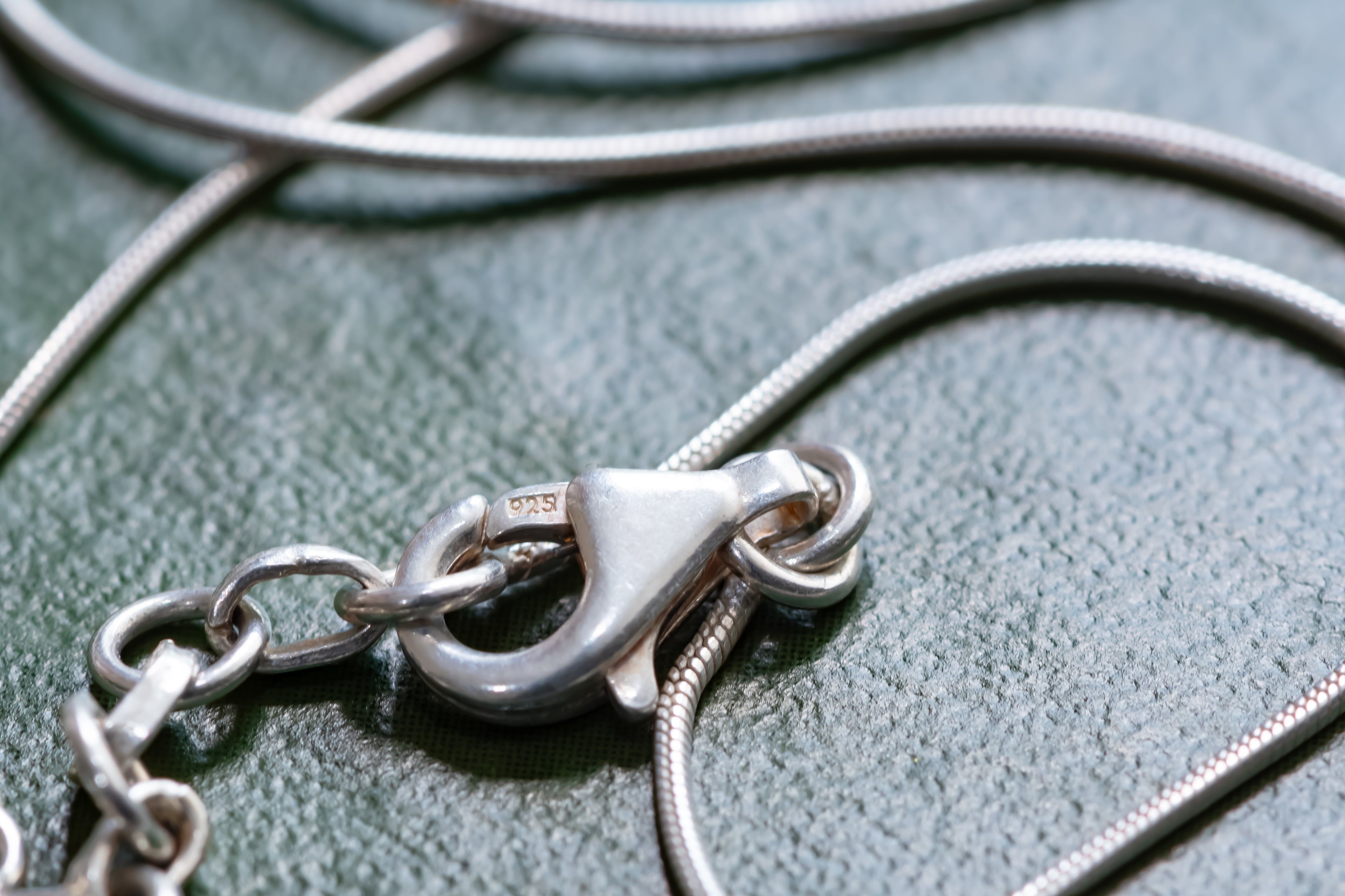Close-up of a silver lobster clasp on a delicate silver snake chain necklace, resting on a textured dark green surface.