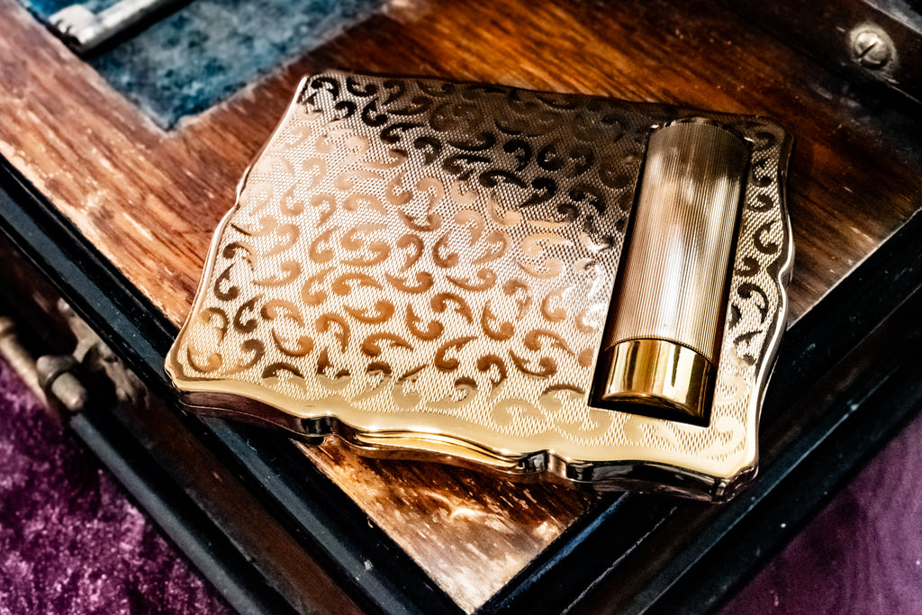 A close-up of an ornate gold-toned metal case with engraved scrollwork and a cylindrical amber metal tube inserted into one corner, resting on a dark wooden surface.