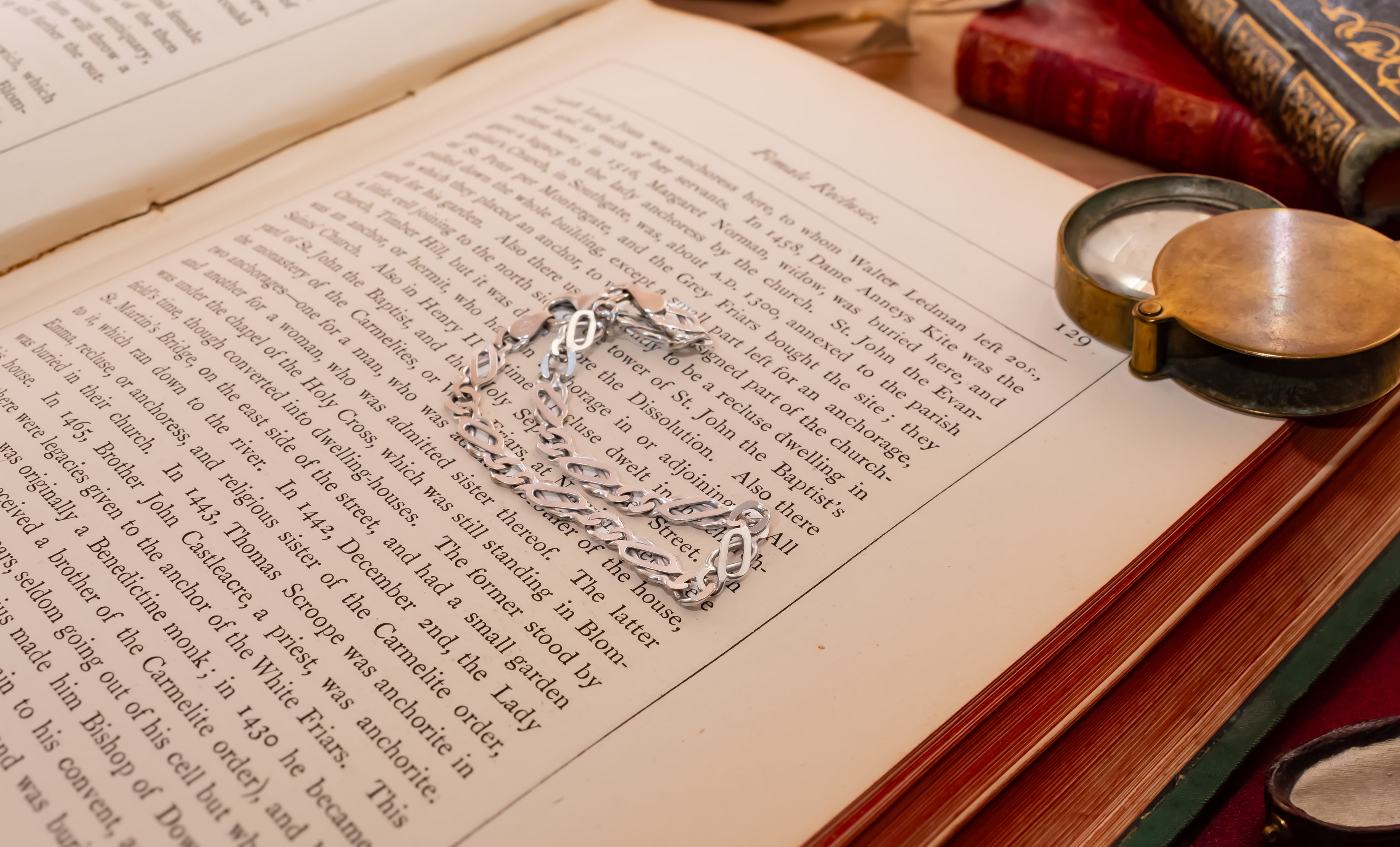 A silver-toned chain bracelet with a small pendant rests on the open pages of an antique book, surrounded by old books and a brass pocket mirror on a wooden surface.