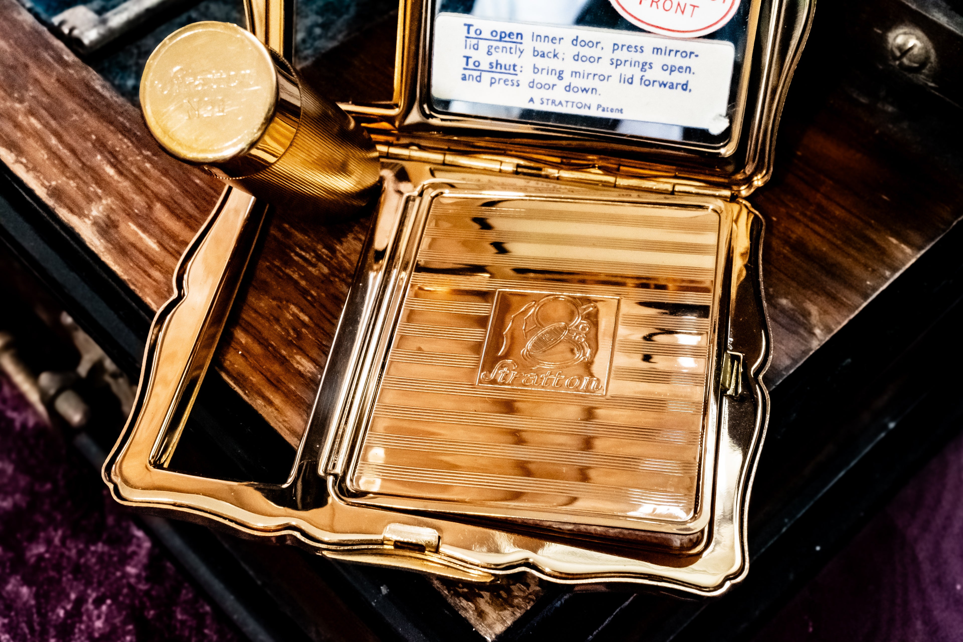 An ornate gold-toned pocket mirror case shown open, with a mirror, inner lid, and engraved interior panel. A small instruction label is visible inside the lid, and the case is photographed close up on a dark wooden surface.
