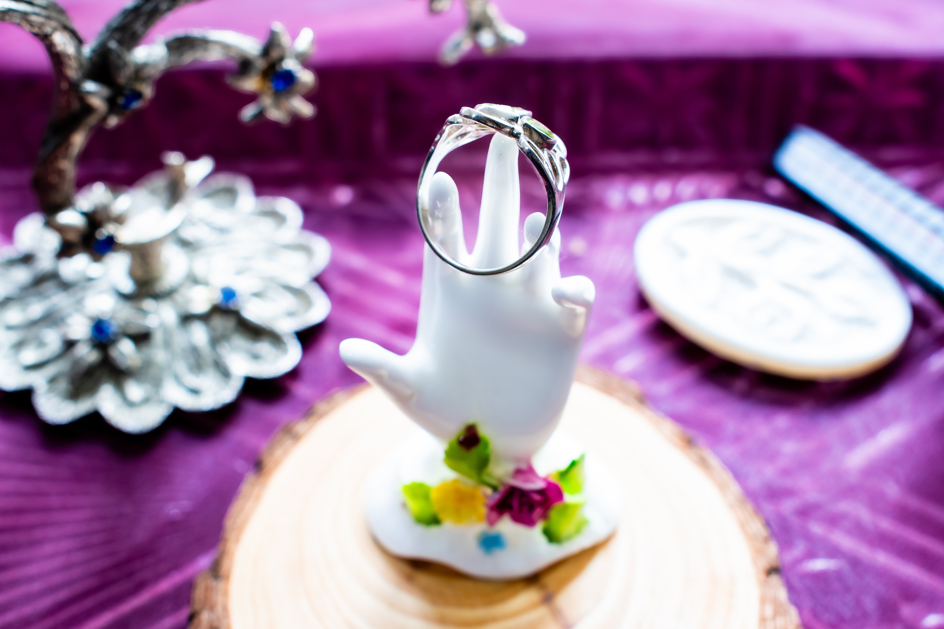 A silver band ring displayed on a white hand-shaped ring holder, set on a small floral base atop a round wood slice, with a purple textured background and blurred decorative objects around it.