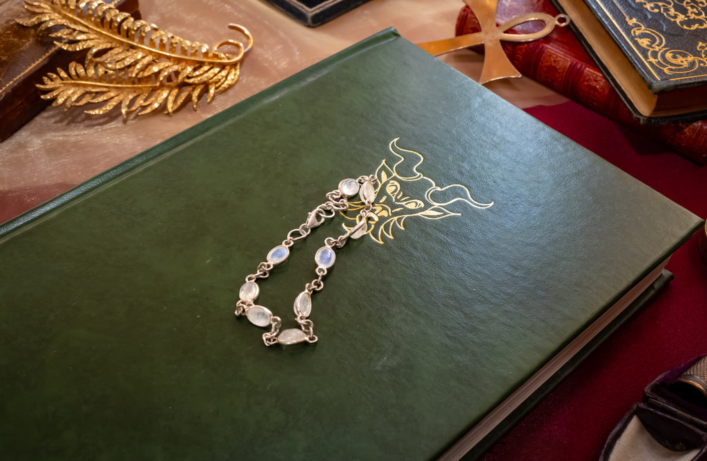 A silver-toned chain bracelet with oval iridescent moonstone-like beads rests on the cover of a dark green hardbound book. The book has gold foil artwork on the front and is photographed in a styled tabletop setting with other books and decorative objects around it.