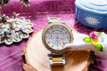 A Michael Kors watch with a gold and silver-tone metal bracelet, a crystal-embellished bezel, and a white dial with gold details, displayed on a wooden slice with decorative props.